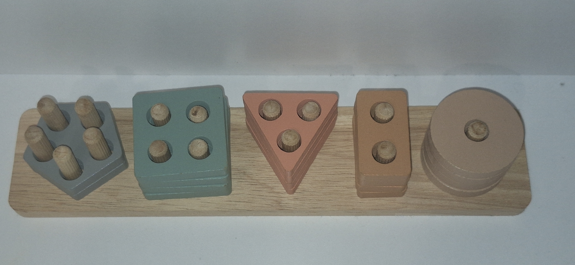 The image shows a children's wooden educational toy on a white background. The toy consists of a light-colored wooden base with five different shaped blocks mounted on it. From left to right, the blocks are: a grey square with five wooden pegs, a teal square with four wooden pegs, a coral triangle with three wooden pegs, a brown rectangle with two wooden pegs, and a stack of beige rings with a central hole. The blocks appear to be designed for sorting and stacking. There is no information in the image to indicate its location in Bila Tserkva, Ukraine.
