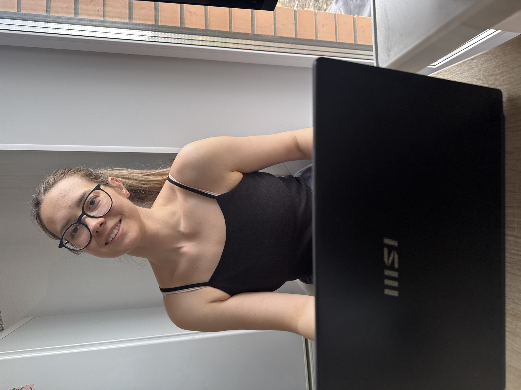The media file presents a portrait of a young woman smiling brightly at the viewer. She has light brown hair, wears black-rimmed glasses, and is dressed in a black spaghetti-strap tank top, with a hint of a lighter strap underneath. The image is captured from a low angle, and appears rotated, with the woman looking towards the camera from the left side of the frame. To her right, a black laptop with the "MSI" logo prominently displayed on its lid occupies a significant portion of the image. The woman appears to be seated or leaning over a white surface, possibly a desk or counter. In the upper part of the background, outside a window or door frame, part of a reddish-brown brick wall is visible, suggesting an exterior view from an indoor location. The presence of natural light indicates it is daytime. The woman's positive expression conveys a happy or content mood. The scene is set in Chaikovsky, Russia.
