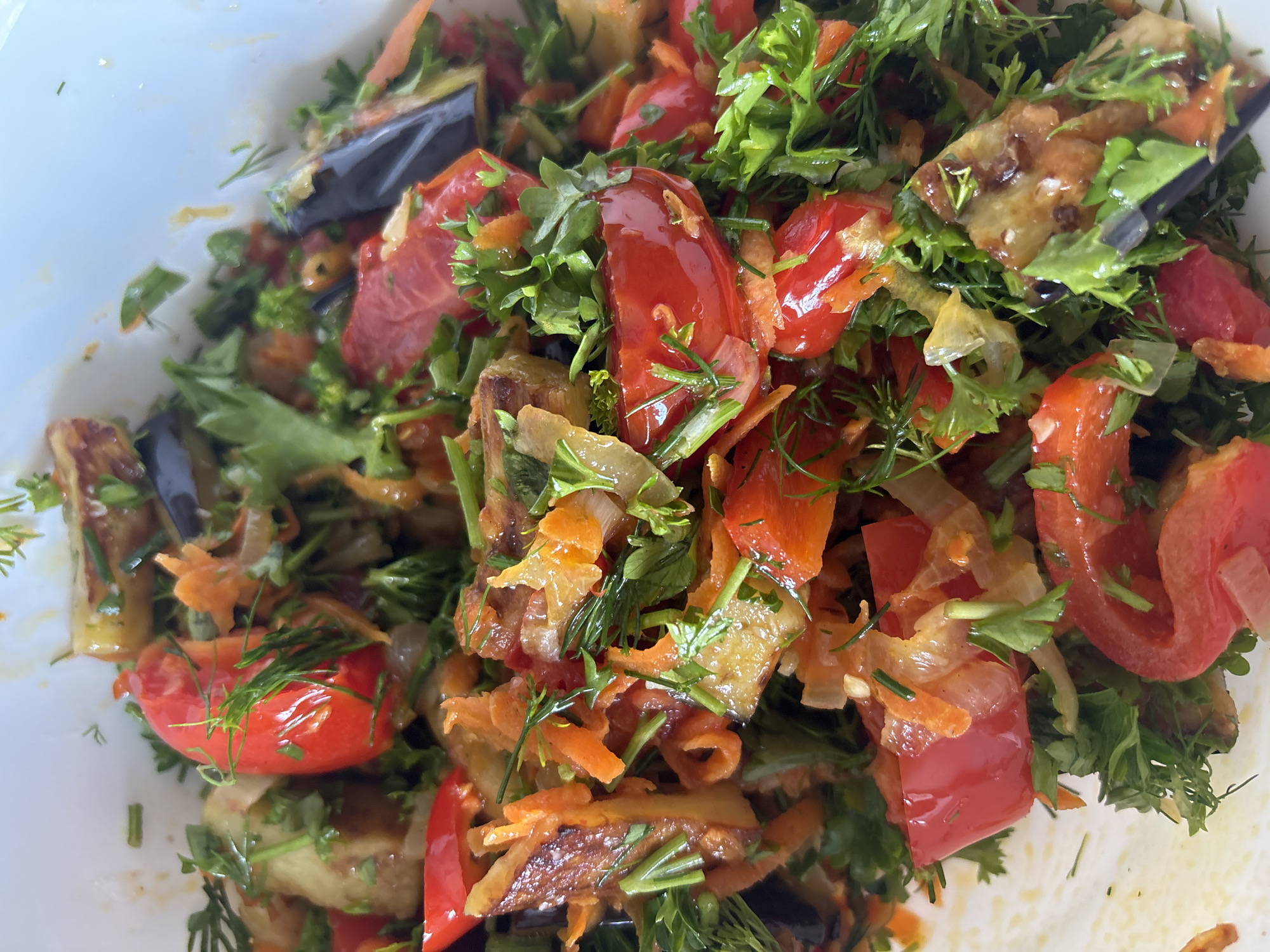 The image shows a close-up, overhead view of a dish containing a colorful mix of chopped vegetables and herbs, likely a salad or a cooked side dish. Dominant ingredients include bright red pieces of tomato, thinly sliced orange carrots, dark purple strips of eggplant, and a generous amount of fresh green herbs, possibly parsley and dill. Some translucent white pieces, likely onions, are also visible. The vegetables appear to be glistening, suggesting they might be dressed or recently cooked.
The scene is tightly cropped on the food itself, presented in what appears to be a white bowl or container, with the edge of the container visible on the left and bottom of the image. The lighting suggests an indoor setting, possibly a kitchen or dining area, with natural light illuminating the dish. The focus is entirely on the food, creating an appetizing visual. There is no text visible in the image. The context provided suggests the location is Bila Tserkva, Ukraine, but this cannot be determined from the image itself.