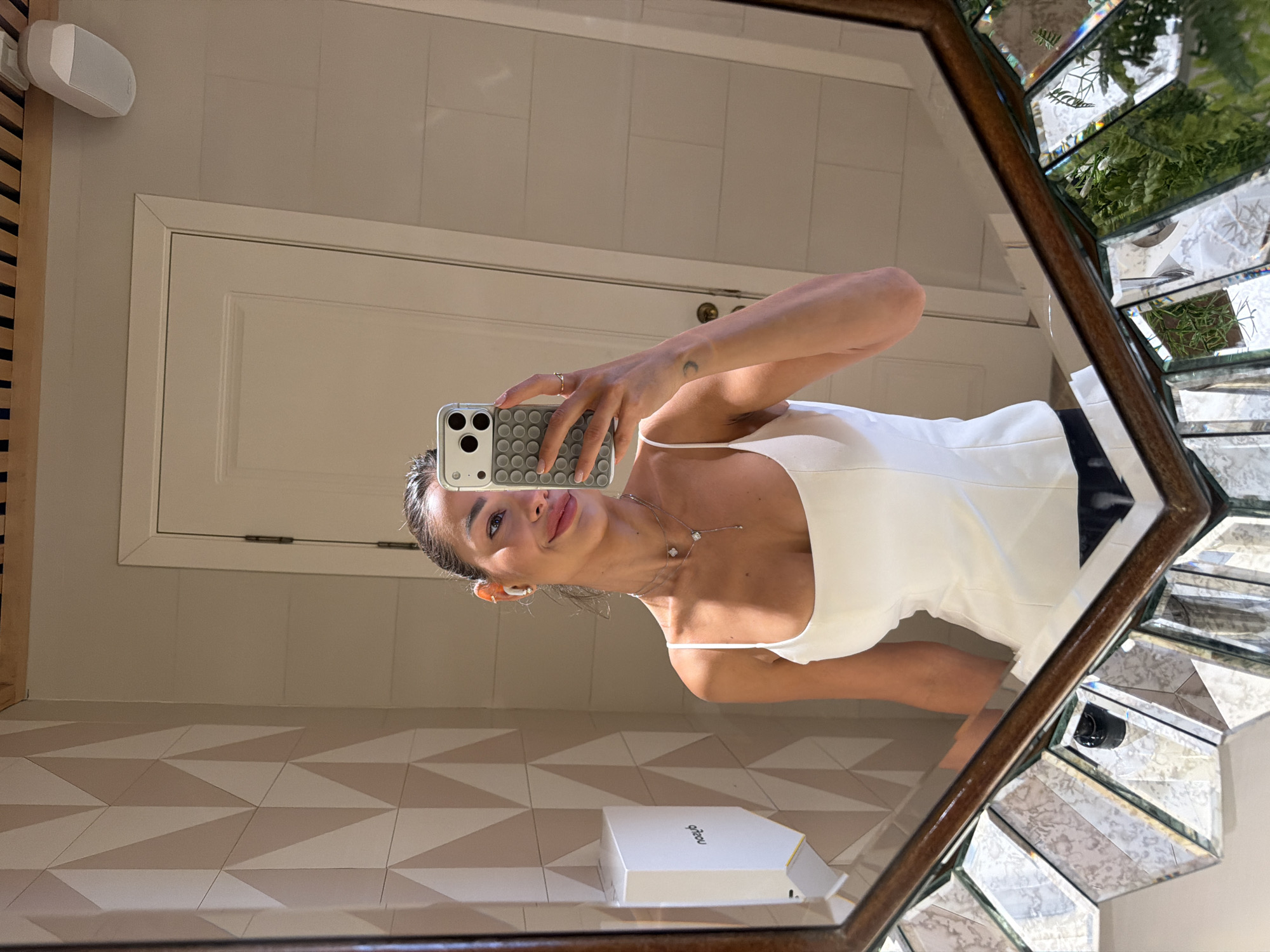 An eye-level mirror selfie captures a woman holding a light-colored smartphone with a gray textured circular-dimpled case in her right hand. She has dark hair pulled back, is wearing white wireless earbuds, and displays a subtle smile while looking towards the phone's camera. She wears a white, fitted, spaghetti-strap top and multiple delicate silver necklaces, including a lariat style. A small crescent moon tattoo is visible on her left forearm, and a gold ring is on her right ring finger.
The background, visible in the reflection, features a bathroom-like setting. The wall on the left has light and dark beige geometric triangular tiles. A white, wall-mounted dispenser with the reversed word "noub" is affixed to this tiled wall. The wall on the right is covered in horizontally laid, rectangular light beige tiles, with a plain lighter wall section above them. A white paneled door with hinges is reflected on the far right. High on the right wall, an oval white speaker is mounted. The ceiling above the door is composed of parallel wooden slats. The mirror itself has a dark, ornate wooden frame and a lower border of numerous beveled, faceted mirror panels that reflect ambient light and what appears to be green foliage from below or to the side.
There are no discernible elements within the image that identify the location as Moscow, Russia.