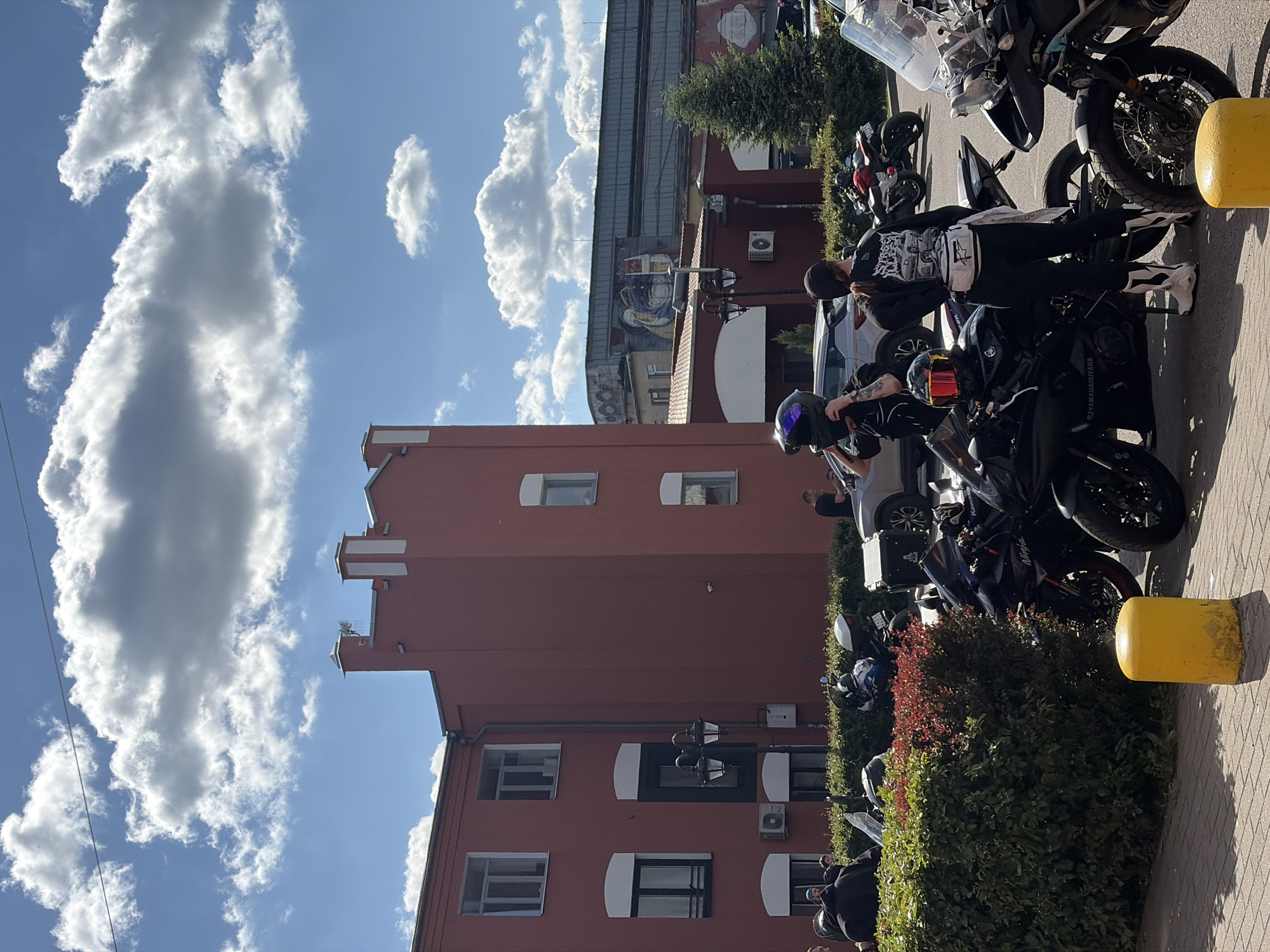 The image shows a street scene in Minsk, Belarus. A red building with white window frames and a distinctive castellated roofline dominates the background. A group of people, some wearing motorcycle helmets and attire, are gathered around several motorcycles parked along a sidewalk. A yellow bollard marks the edge of the parking area. The sky is blue with scattered white clouds. There are plants and trees near the building.