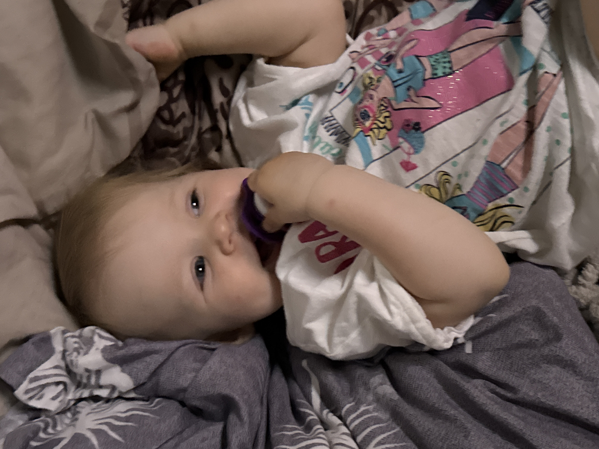 A baby lies on a bed covered with patterned sheets and blankets. The baby has light skin, blue eyes, and blonde hair. The baby is sucking on a purple pacifier and wearing a white shirt with a graphic design of cartoon figures. The location is not specified in the image and cannot be determined from the provided details.