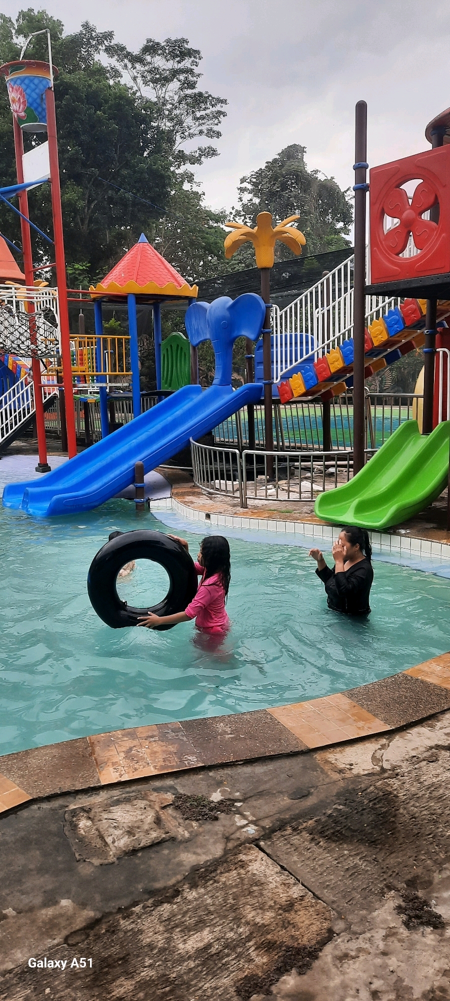A family is enjoying a day at the aquatic playground in Popoh, Indonesia. Two young girls are in the shallow end of the pool. One girl, wearing a pink shirt and swim bottoms, is holding a black inflatable ring. The other girl, wearing a black long-sleeved shirt and swim bottoms, is standing with her hands covering her face. In the background, a large blue slide curves into the pool, and beyond that, a red and blue playground structure with multiple slides and climbing areas is visible. Trees and a cloudy sky form the backdrop. The pool is surrounded by a tiled deck and a metal fence. The ground surrounding the pool is made of concrete with some patches of dirt and vegetation. The water is clear and blue.