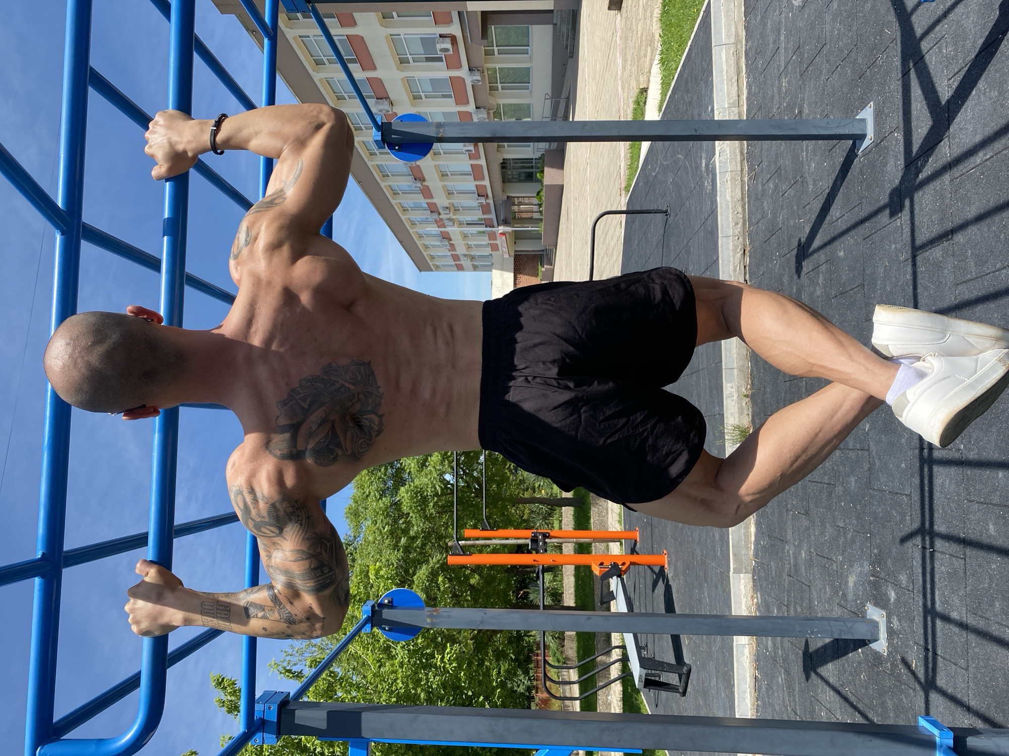 A muscular, bald man with tattoos is suspended horizontally between two parallel blue bars at an outdoor fitness park. He wears black shorts and white sneakers. The fitness park includes gray and blue metal structures on a dark gray rubberized surface. In the background, there is a multi-story residential building with a light-colored facade. Trees and greenery are also visible. The image was taken in Pleven, Bulgaria.