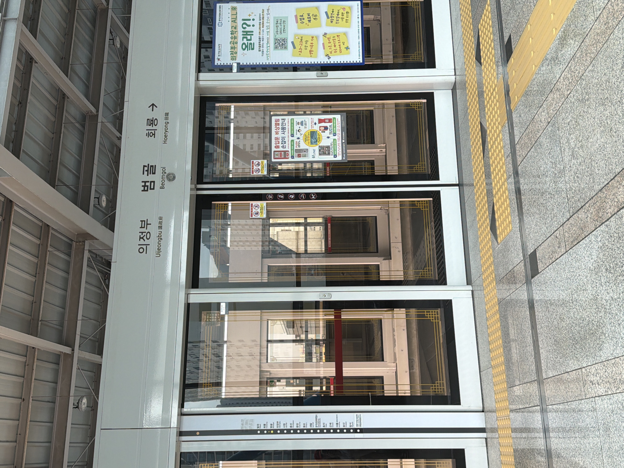 The image shows a subway platform in Uijeongbu-si, South Korea. The platform has automatic doors that are closed, revealing the interior of the subway cars through the glass. Above the doors, a sign displays station names in Korean and English: "의정부 (Uijeongbu)", "범골 (Beomgol)", and "회룡 (Hoeryong)" with an arrow pointing to the right. There are yellow tactile paving strips on the ground in front of the doors. A poster with Korean text is visible on the right side of the image, likely advertising for the "Uijeongbu Education ALL" program. The environment appears clean and modern, typical of a public transportation hub.