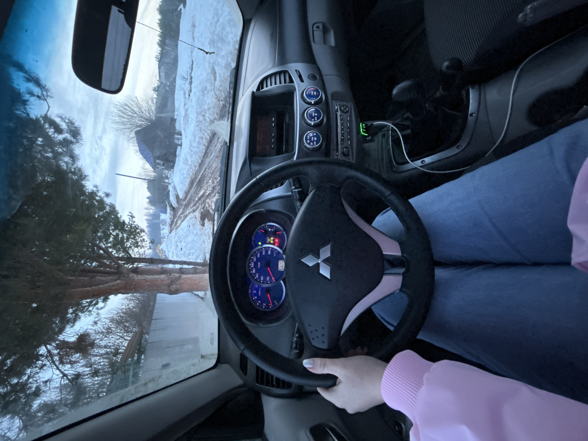 The image depicts the interior of a vehicle, specifically from the driver's perspective, looking out onto a rural scene in Фоки, Russia. The driver's left hand, with white nail polish, grips the black steering wheel, which features a Mitsubishi logo. The driver wears a light pink sweatshirt and blue jeans.
The dashboard displays blue-lit gauges for speed, RPM, fuel, and temperature. The center console includes an infotainment screen, three rotary climate control knobs, and a stereo unit. A green-glowing USB charger with a white cable is plugged into a power outlet. A manual gear stick and handbrake are visible to the right of the driver's leg. A rearview mirror hangs from the ceiling, reflecting the sky, with a small decorative item suspended from it.
Outside the vehicle, a muddy dirt road is partially covered in snow. Snow is also present on the verges and in distant fields. To the left, several evergreen trees stand beside a corrugated metal fence. In the mid-ground, bare deciduous trees are visible, along with a small, weathered house featuring a blue roof. Further right, a pile of snow-covered material is present. The sky is overcast with patches of brighter light.