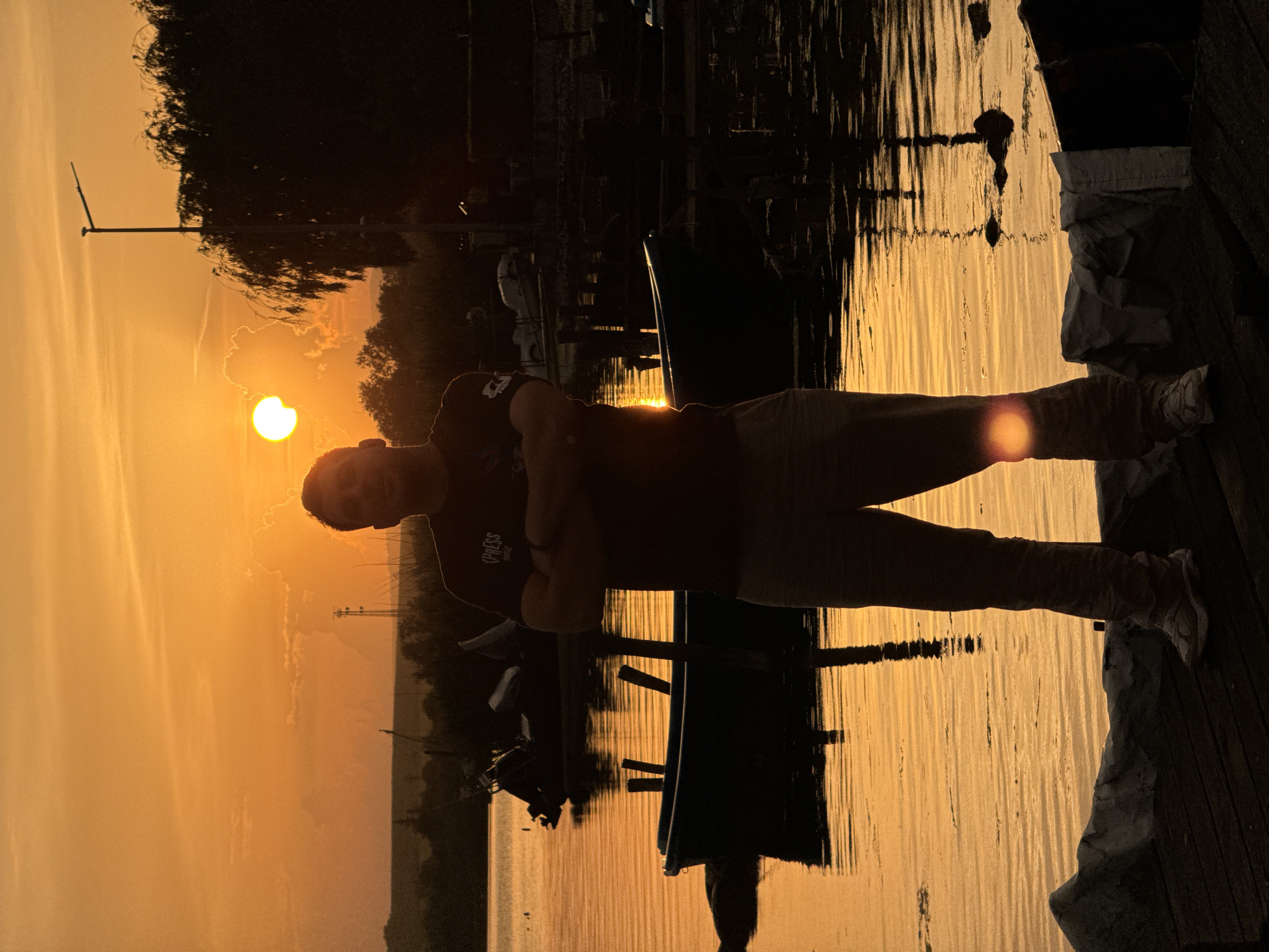 A man is standing on a wooden dock next to a body of water at sunset. He is wearing a dark t-shirt, light-colored pants, and sneakers. The sun is visible on the horizon and reflecting on the water. There are boats and trees in the background. The image was taken in Казашко, Bulgaria.