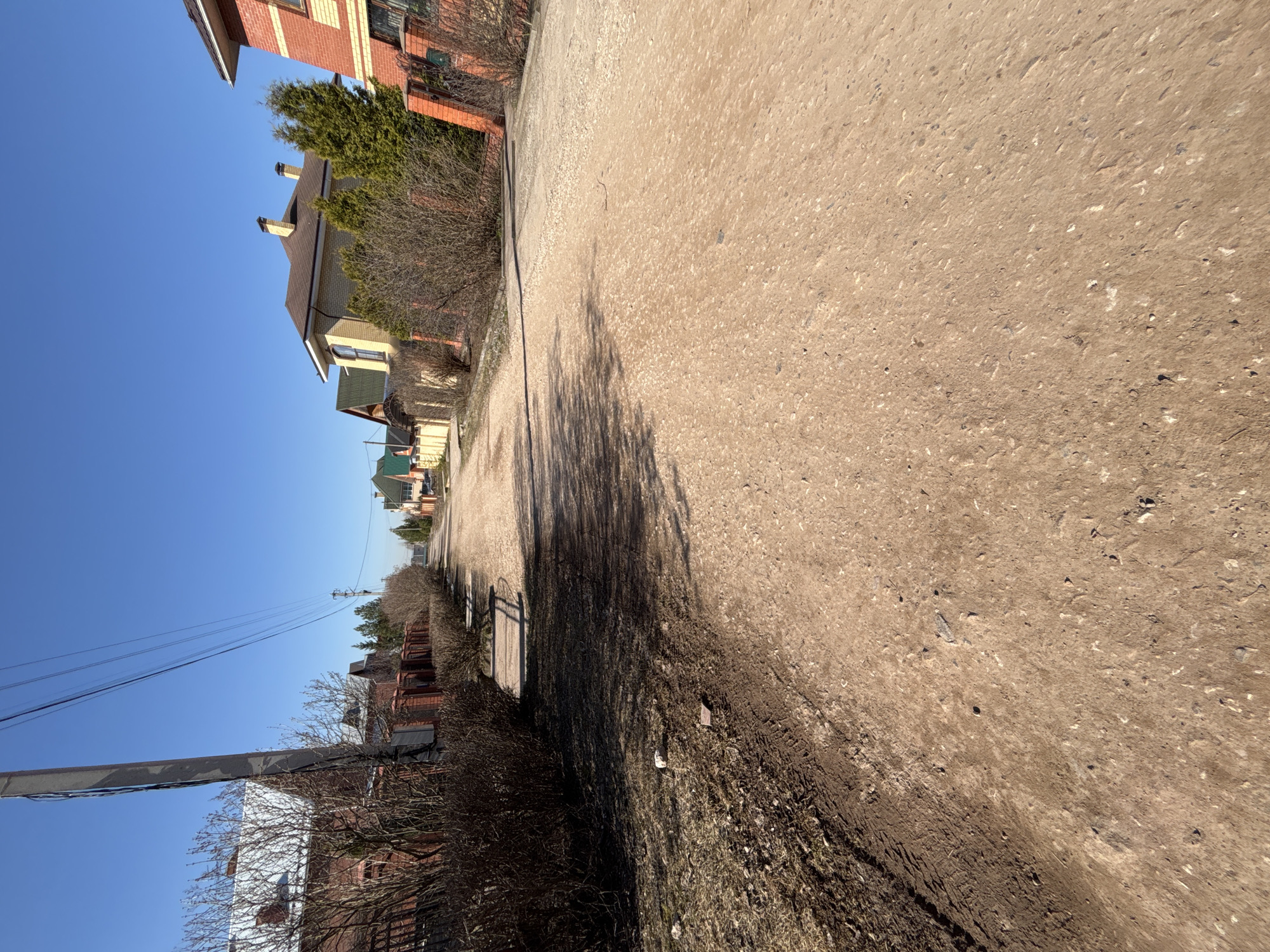 The image depicts a street scene in Ферма, Russia, oriented at a slight angle. The road, made of dirt or gravel, dominates the foreground and extends into the distance. Along the left side of the road are various houses, some made of brick and others with light-colored siding. A dense hedge runs alongside the road, casting a significant shadow onto the road surface. A utility pole is visible on the left, with overhead power lines extending along the street. The sky is clear and blue.