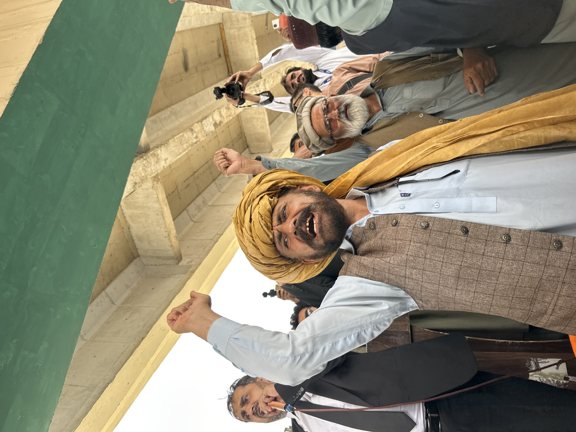 A group of men are gathered outdoors, under a green and beige overpass. The primary subject, a man in the foreground, wears a yellow turban, a light blue shalwar kameez, and a brown checkered waistcoat. He has a beard and mustache and his mouth is open as if speaking or shouting. His right arm is raised with his fist clenched. To his left, a man in a black suit and tie, also with a microphone, appears to be speaking. Behind them, other men are visible, some holding cameras and one holding a video camera above his head. The overall atmosphere suggests a political rally or protest, potentially in a public area of Quetta City Tehsil, Pakistan, given the visual context of the men's attire and the presence of a microphone and cameras.