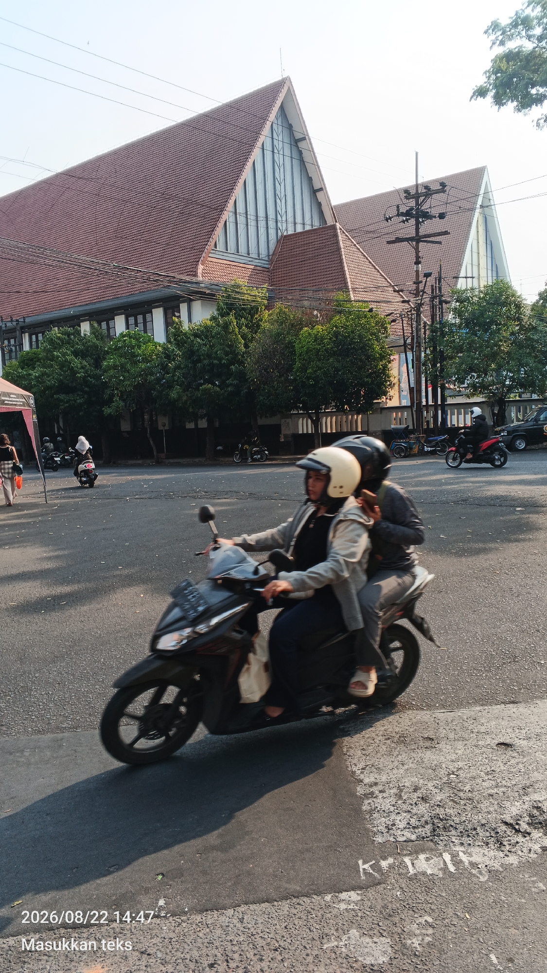 The image shows a street scene in Malang, Indonesia, with two people riding a motorcycle in the foreground. The woman in front is wearing a white helmet and a grey jacket over a black shirt. The man behind her is wearing a grey long-sleeved shirt and has a dark helmet. They are both looking forward, and the motorcycle is in motion. In the background, there are two large buildings with distinctive triangular roofs, likely a church or a civic building, with trees and power lines in between. Other motorcycles and cars are visible in the distance on the street. The lighting suggests it is late afternoon. The timestamp in the lower left corner indicates the image was taken on August 22, 2026, at 14:47.