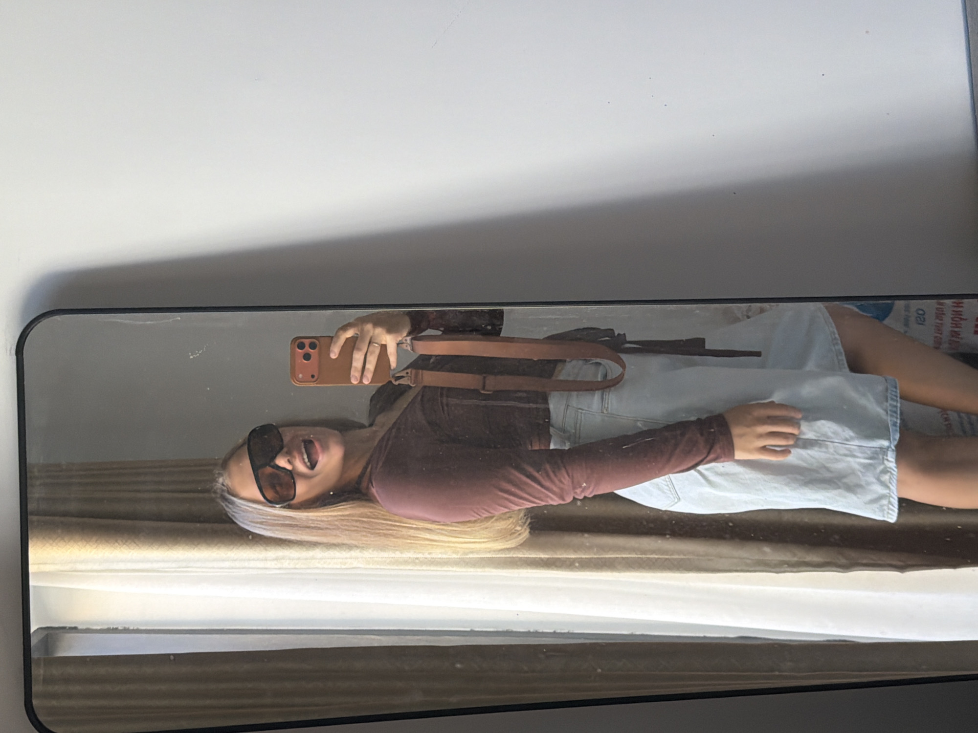 A young woman with blonde hair is standing in front of a mirror, taking a selfie. She is wearing large, dark sunglasses, a long-sleeved brown shirt, and a light blue denim skirt. The mirror reflects her and a window with sheer curtains in the background. The wall to the right of the mirror is plain white, with a shadow cast on it from the side. The image appears to be taken indoors, likely in a residential setting, based on the presence of the mirror, curtains, and a white wall. No specific landmarks or indicators of Bac Nha Trang Ward, Vietnam are present in the image.
