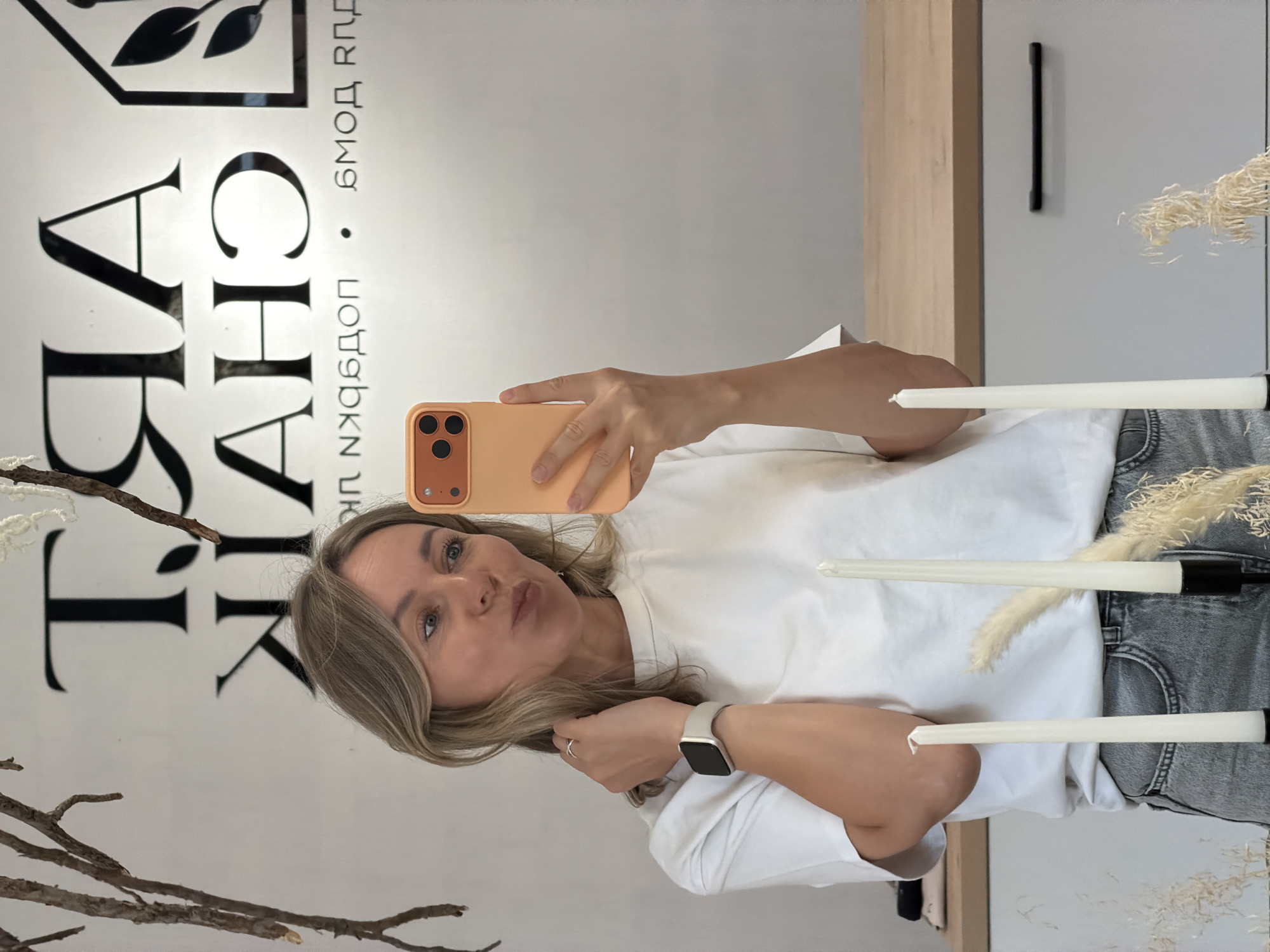 A woman is taking a selfie in a mirror. She is wearing a white t-shirt and grey jeans, with a smartwatch on her left wrist. She is holding an orange phone in her right hand. In the foreground, there are three white taper candles in candleholders and some dried grass. In the background, there is a sign that says "ART CHAIK". The woman is in Chaikovsky, Russia.