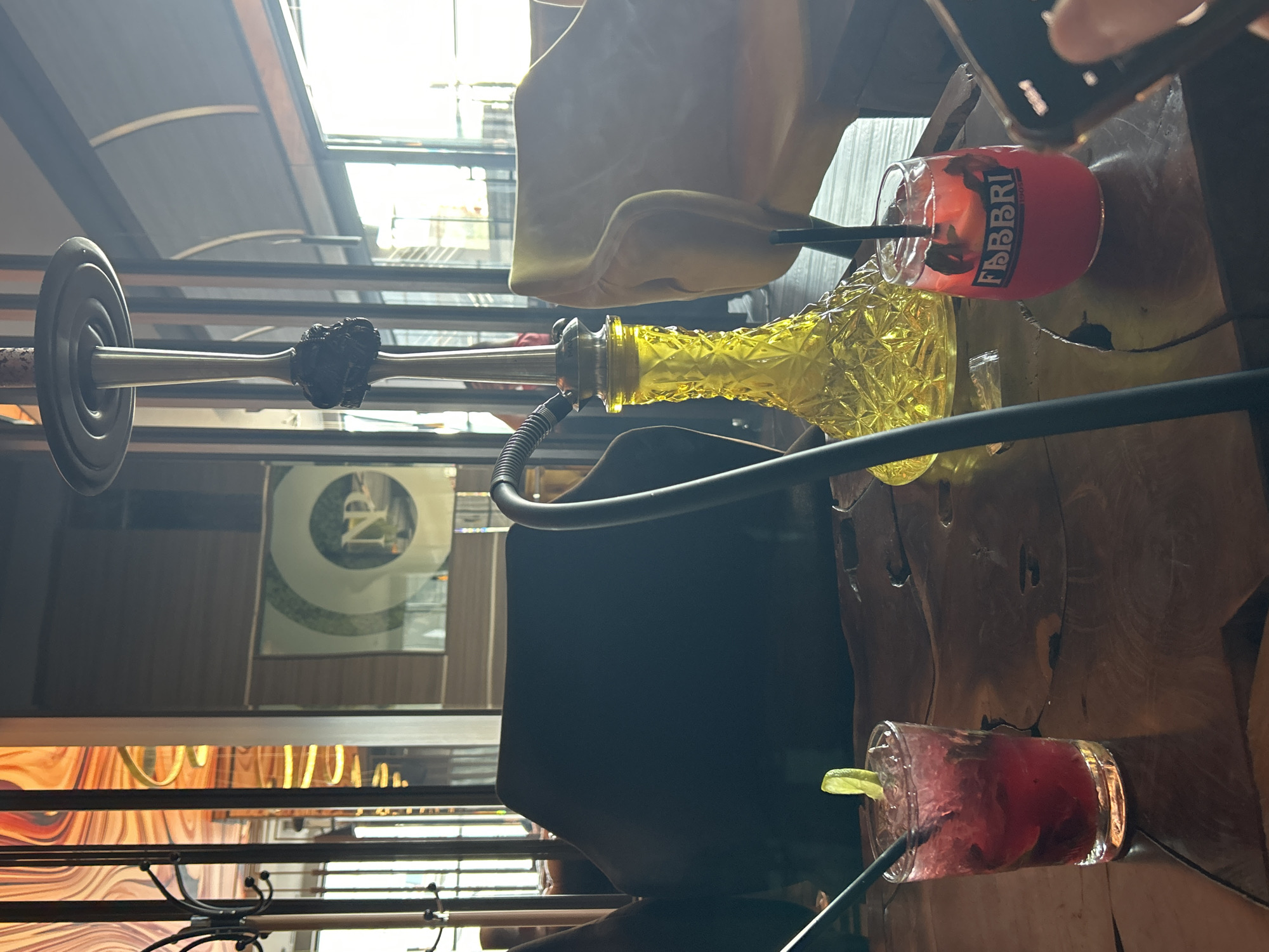 The image captures a vibrant and relaxed social setting, likely a cafe or lounge in Burgas, Bulgaria, illuminated by natural daylight. On a dark wooden table, two red-hued beverages and a yellow, textured glass hookah are prominently displayed.
One of the red drinks, served in a clear glass with ice, distinctly features the vertical text "FABBRI 1905" on its side, indicating a brand. The other red drink, also in a clear glass with ice, is garnished with a fresh lime wedge. A yellow glass hookah, intricately patterned, stands between the drinks, featuring a metal stem and a black hose.
In the upper right corner, a person's hand holding a dark-colored smartphone is partially visible, suggesting candid photography or a moment of engagement. The background reveals a light brown upholstered chair or cushion, and bright daylight streams through large windows or a skylight, casting reflections and showing hints of building structures. On a dark surface in the mid-background, reflections show abstract patterns and a circular logo with stylized letters, possibly "NP".
The scene depicts individuals enjoying refreshments and a hookah in a comfortable, modern environment, creating an atmosphere of leisure and socializing during the daytime.