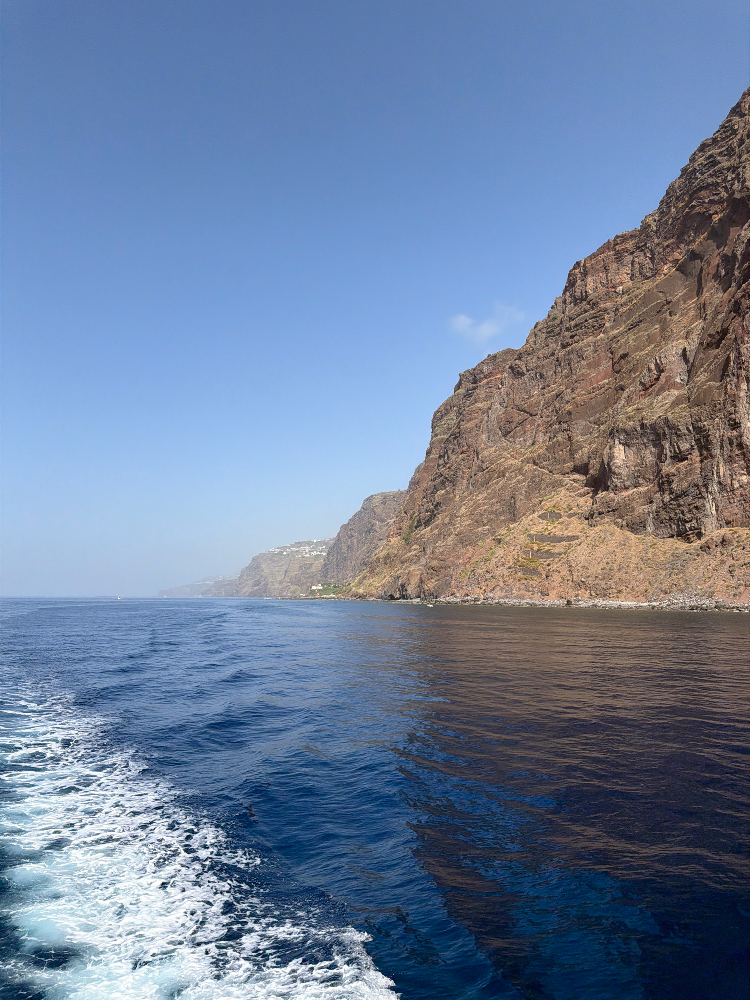 This is a photograph taken from a boat looking towards the coastline of Câmara de Lobos, Portugal. The image features a steep, rocky cliff face on the right side of the frame, with the ocean occupying the left and foreground. The water is a deep blue, with white foam and waves trailing behind the boat, indicating it is in motion. The cliff is rugged and appears to be composed of reddish-brown rock, with some sparse vegetation and signs of human habitation, including a few white buildings clustered on the hillside in the distance. The sky is clear and blue, with a hazy quality towards the horizon. The overall scene depicts a coastal landscape.