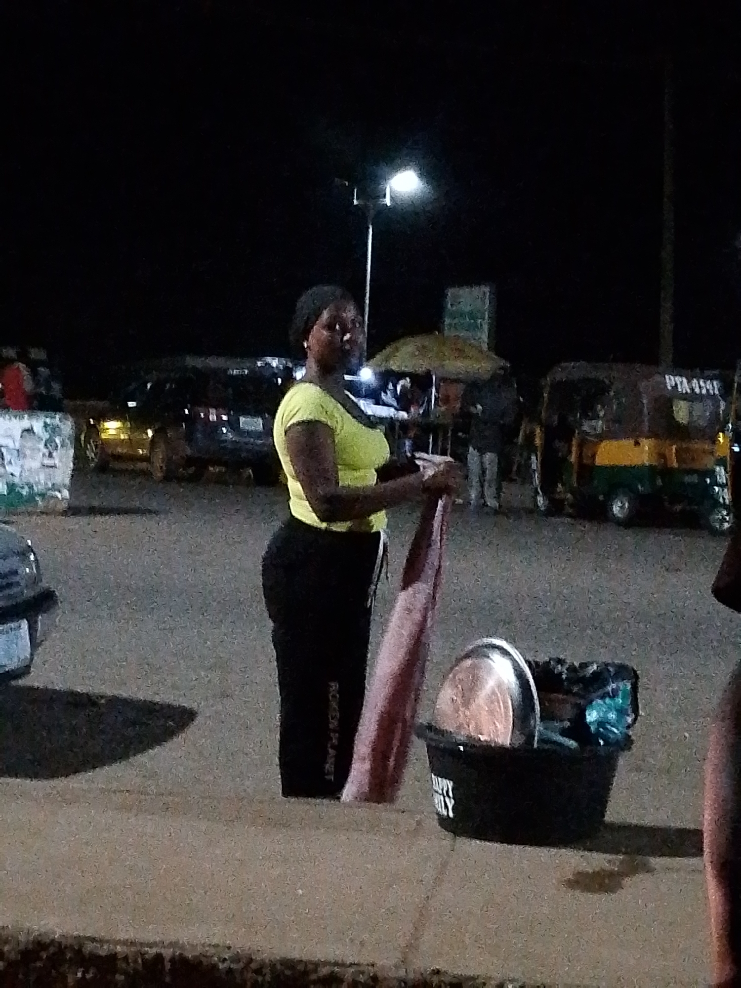 A woman is standing on a sidewalk at night, holding a pink fabric and a metallic pot. She is wearing a lime green top and black pants. In front of her is a black bucket with a silver pot and other items inside. Behind her, there are several vehicles, including a yellow and green auto-rickshaw with the license plate "PTA 454f". The background includes streetlights and other buildings. The scene appears to be a street in an urban area at night.