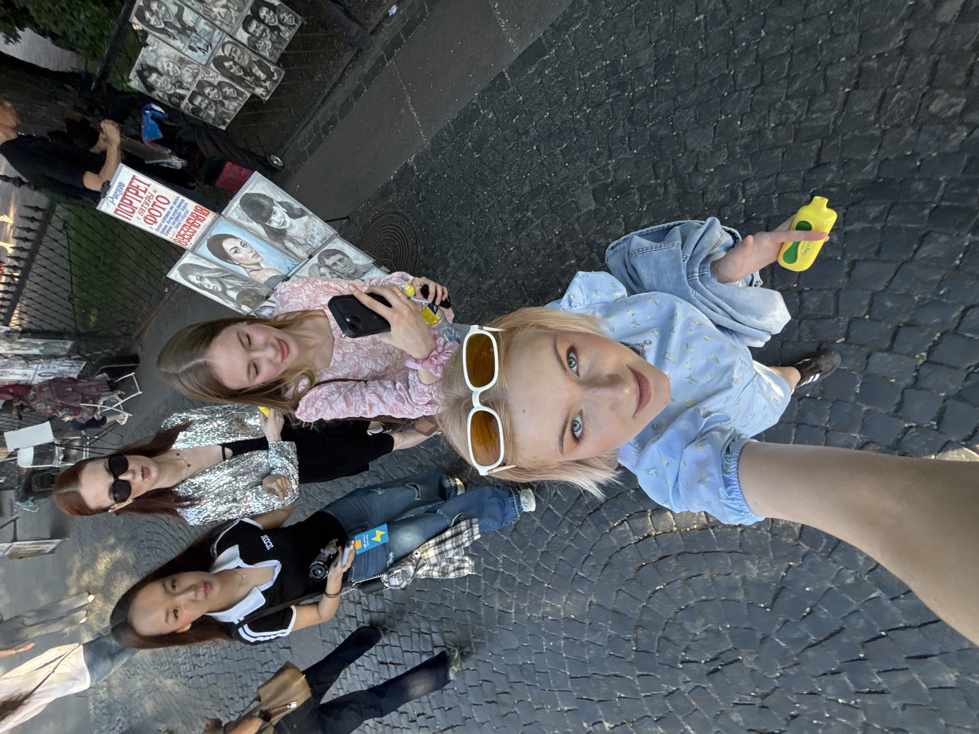 A high-angle selfie captures five young women on a dark cobblestone street in Saint Petersburg, Russia, adjacent to a black metal fence with trees beyond. In the foreground, a blonde woman with blue eyes and white sunglasses pushed onto her head, wearing a light blue floral blouse and light blue jeans, extends her right arm to take the photo while holding a yellow, stoppered bottle in her left hand. To her right, a woman with light brown hair in a pink floral top and dark pants looks down at a black smartphone held in her left hand, also holding a yellow bottle. To the left, a woman with long dark hair wears a black polo shirt with "ICON" text, ripped blue jeans, and white sneakers; a plaid shirt is tied around her waist, and she holds a black camera lens and a smartphone. Next to her, a woman with reddish-brown hair, dark sunglasses, a silver sequined jacket, a black top, and black pants stands with her left hand slightly raised. Partially visible on the far left, another person walks away in blue jeans and a white shirt, carrying a brown bag. In the background, street artists display numerous framed, mostly monochromatic, charcoal or pencil portraits on stands, alongside a white sign with Russian text "Портрет Фото" (Portrait Photo) and "c натуры" (from nature/live) above a phone number. The scene is outdoors under natural light.