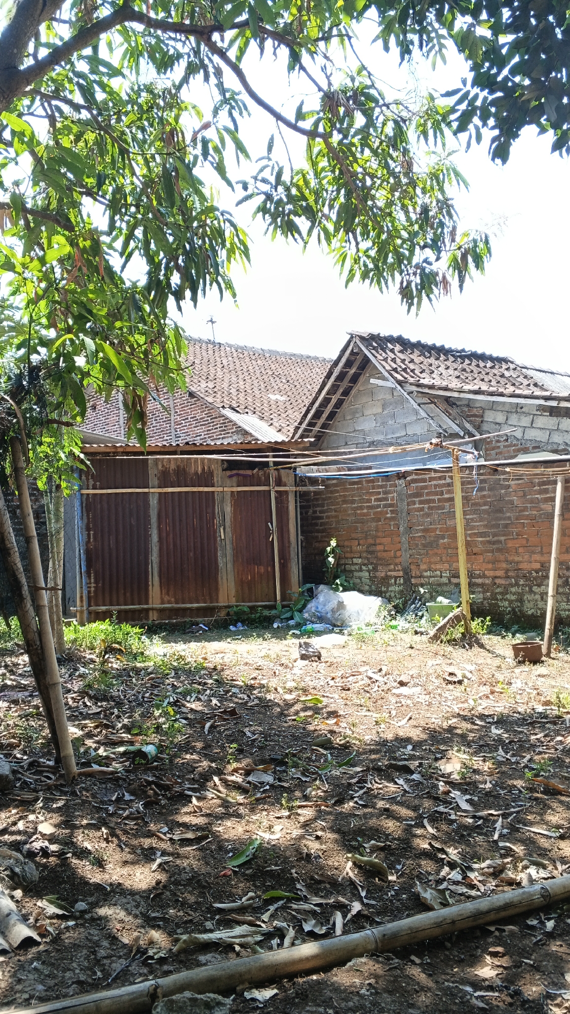 This is a photograph taken in Karangjati, Indonesia, depicting an outdoor scene behind two structures. In the foreground, dry leaves and dirt cover the ground, with several pieces of bamboo scattered horizontally and vertically. Sunlight filters through the branches of a tree on the left, casting shadows on the ground.
Behind the foreground, a rusty corrugated metal gate is partially visible, flanked by bamboo poles and overgrown vegetation. Beyond the gate, a brick wall with a concrete block extension forms the side of a building. The roofs of two structures are visible, both with terracotta tiles. The building on the right features an exposed wooden roof frame. A clothesline with blue and white lines is strung between poles in front of the brick wall. A large white plastic bag and other debris are present on the ground near the wall. The overall impression is of a somewhat unkempt, residential backyard area.