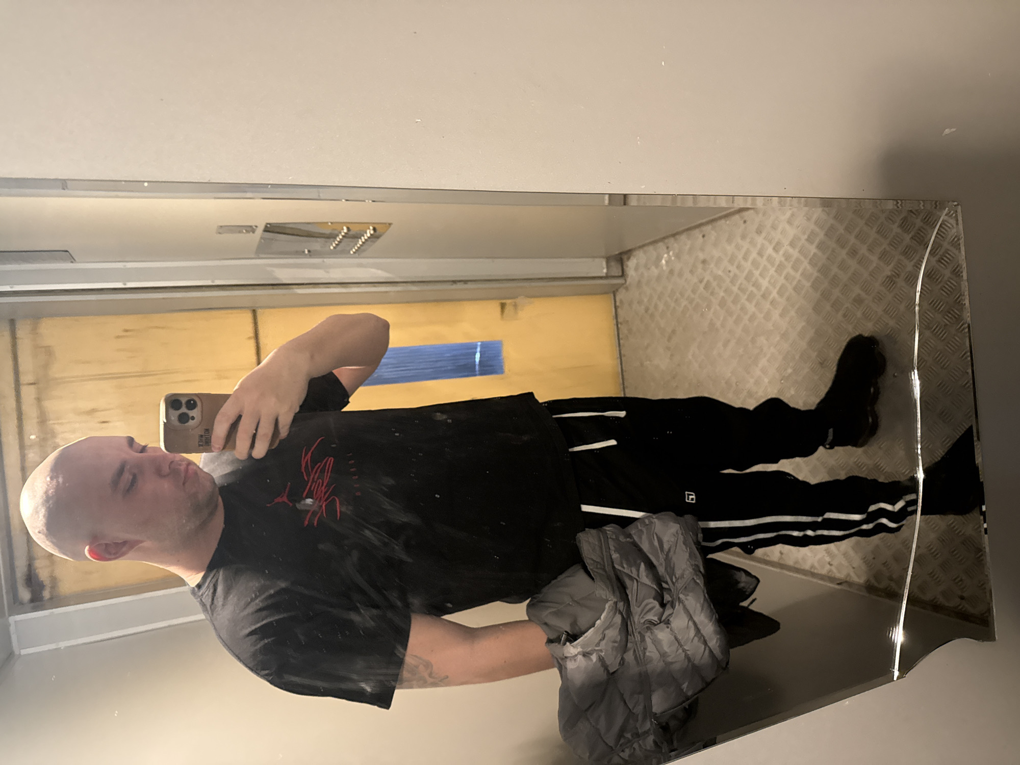 A man is taking a selfie in a mirror. The mirror reflects the man's face, torso, and legs. He is wearing a black t-shirt with a red design, black pants with white stripes, and black shoes. He is holding a smartphone in his right hand. A gray jacket is draped over his left arm. The background is a room with light-colored walls and a diamond-plated floor. A light fixture is visible on the ceiling. The location of the photo is the city of Pleven, Bulgaria.