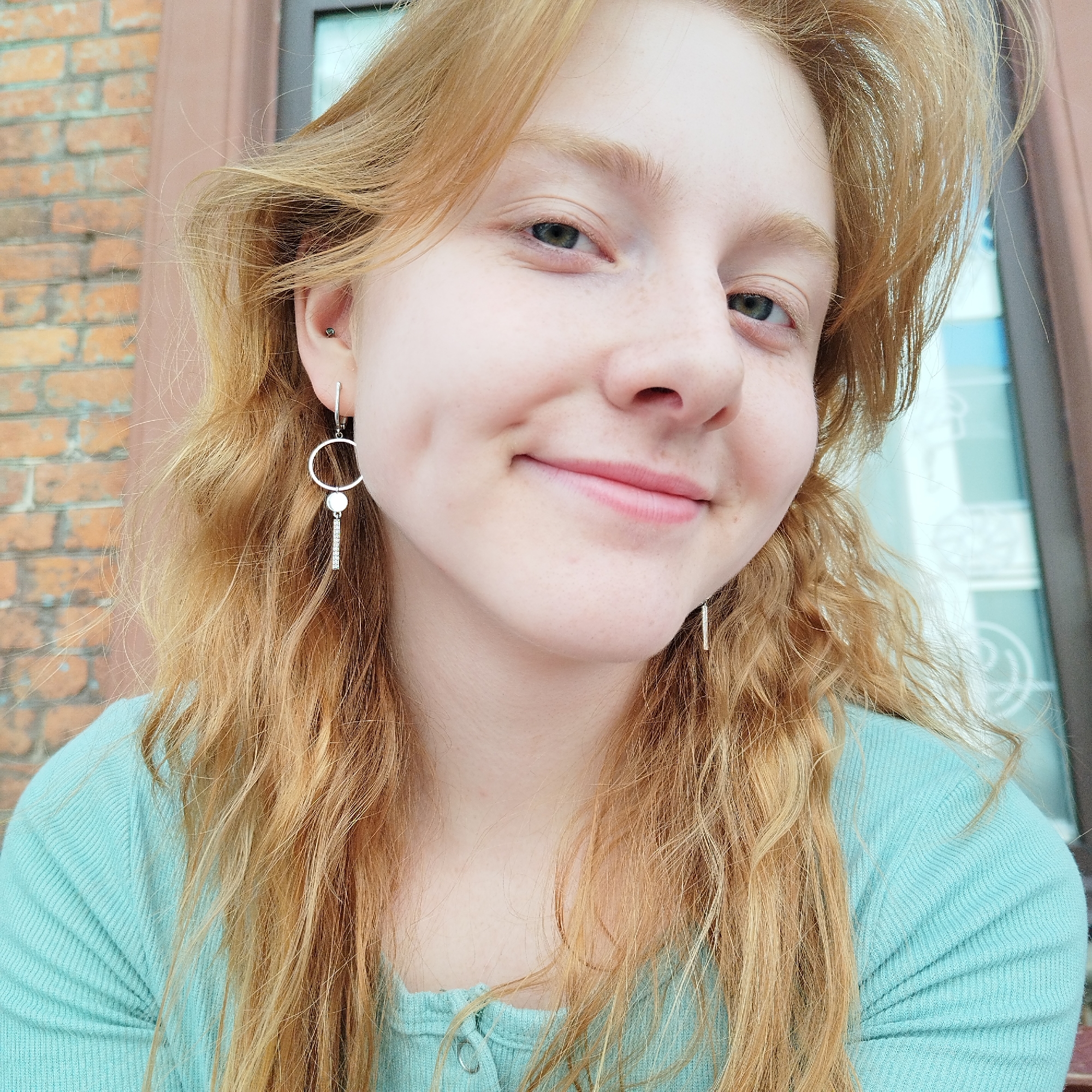 A close-up portrait, captured in natural daylight, features a young woman with fair skin, light red or strawberry blonde hair, and subtle freckles. Her light-colored eyes, possibly green or hazel, gaze directly at the camera as she displays a gentle, closed-mouth smile, creating a dimple on her left cheek. Her wavy hair cascades around her face and shoulders.
She is dressed in a light teal or aqua long-sleeved, ribbed top. In her left ear, she sports a small stud piercing in the upper cartilage and a distinctive dangling earring from the lobe, which comprises a silver hoop, a small circular disc, and a vertical bar. A similar vertical bar earring is visible on the right side of her face.
The background provides context to the setting: a brick wall is prominent on the left, suggesting an outdoor or semi-outdoor location, possibly on a porch or balcony of a building in Yekaterinburg, Russia. To the right, a dark window or doorframe is visible, with a hint of light blue, perhaps sky or a reflective surface. The bright, even illumination indicates the photo was taken during the daytime. The woman's expression conveys a calm and friendly demeanor. No visible text is present in the image.