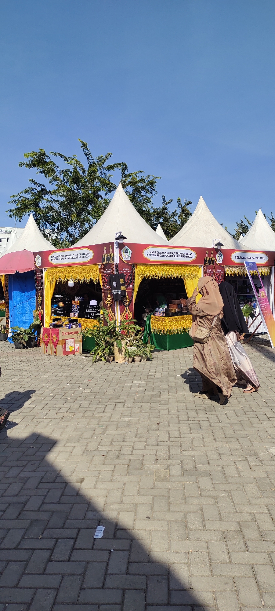 In the city of Meunasahaluedrienlhoksukon Barat, Indonesia, an outdoor market scene unfolds under a clear blue sky. Several white, conical tents are erected, creating vendor stalls. The central tents display banners with Indonesian text, including "DINAS PERDAGANGAN, PERINDUSTRIAN, KOPERASI DAN USAHA KECIL MENENGAH" and "SEKRETARIAT BAITUL MAL," indicating government or business-related entities. Yellow drapes with intricate patterns adorn the front of these stalls, which are filled with merchandise, including what appear to be hats or caps arranged on shelves.
In the foreground, two women are walking away from the camera, towards the right. The woman on the left wears a light brown hijab and a patterned brown dress. The woman on the right wears a black hijab and a light-colored, flowing skirt and top. A light-colored handbag is slung across the left woman's shoulder. The ground is paved with light gray interlocking bricks. To the left of the tents, a blue tarp is visible. Several potted plants are placed around the stalls. In the distance, green trees and a building with white walls are visible. Shadows cast by the tents and potentially other unseen structures stretch across the brick pavement.