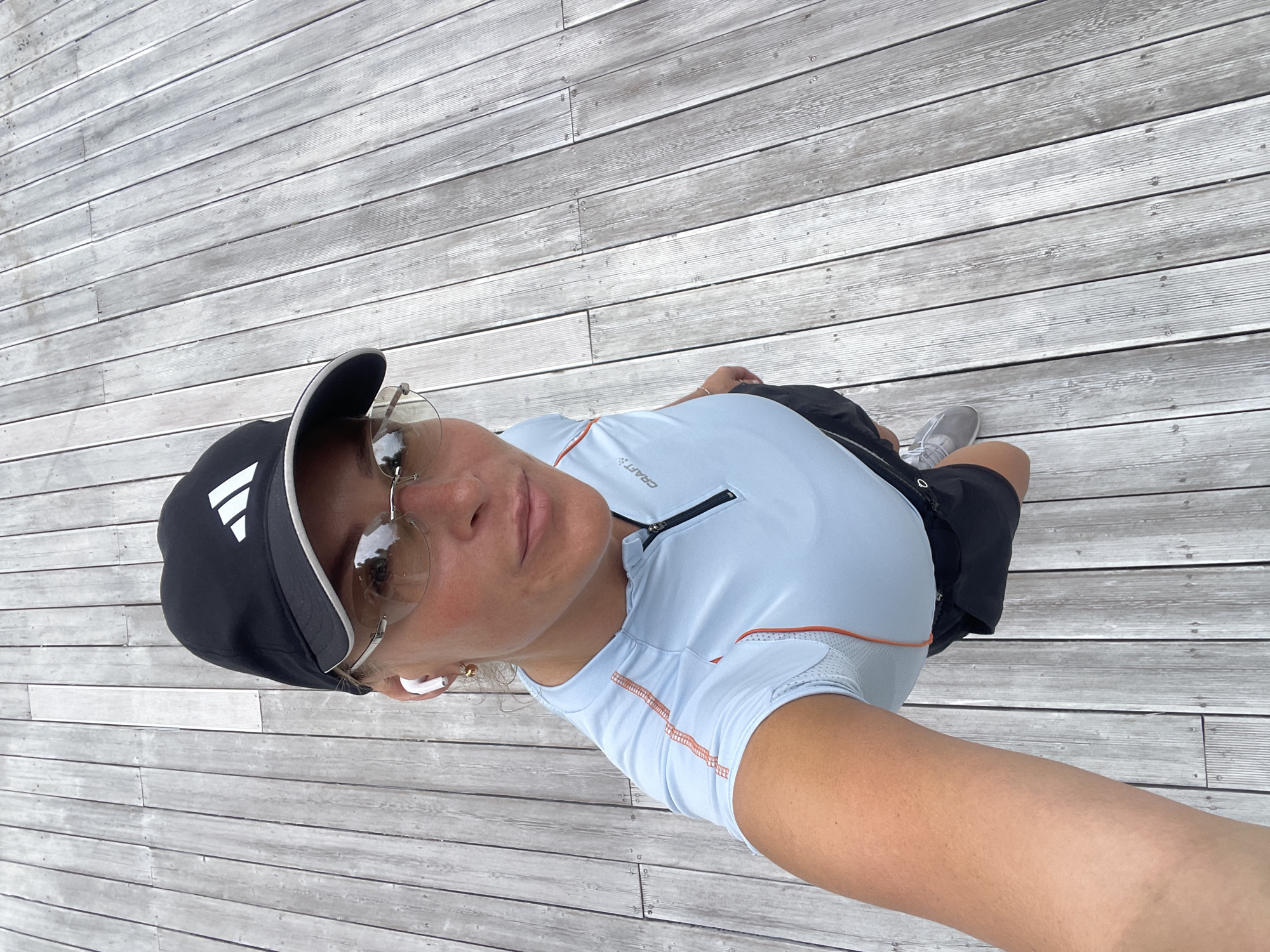 A woman is standing on a grey wooden deck, wearing a black Adidas cap, clear-framed glasses, and a light blue athletic shirt with orange trim. She has an earpiece in her left ear. She is looking up and slightly to the left, taking a selfie with her arm extended. She is wearing black shorts and grey sneakers. The wooden deck has parallel planks running horizontally in the image, and appears to be outside. The image appears to be taken from a high angle. The background consists of the grey wooden deck. There is no visible indication that the photo was taken in Sochi, Russia.