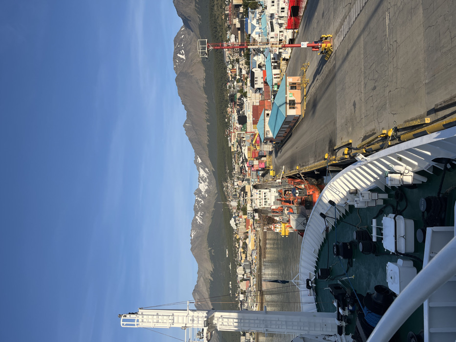 The image captures a detailed view of the port of Ushuaia, Argentina, under a clear, bright blue sky with faint high-altitude cloud streaks. The perspective is from the deck of a large vessel, looking towards the city and its surrounding mountains.
In the immediate foreground, the green deck and white railing of the viewing vessel are visible, along with a prominent white mast structure on the left. Mooring bollards, coiled lines, and white utility boxes are present on the deck.
The midground features a concrete pier where multiple vessels are docked. Two orange-hulled tugboats or workboats are alongside the pier, with one individual wearing a blue shirt visible standing on the stern deck of the closer orange vessel. Further along the pier, a white-hulled vessel is also docked. A light-colored building with a distinct turquoise-colored roof stands on the pier, adjacent to a paved roadway. Beyond this, an area with numerous stacked colorful shipping containers, including some with "Maersk" branding, is visible, indicating cargo operations. A red and white striped barrier arm and a tall red navigational or port signaling pole are positioned on the right side of the pier.
The city of Ushuaia occupies the background, spreading from the waterfront up the lower slopes of the mountains. Numerous buildings of various sizes and colors are densely packed throughout the cityscape. The far background is dominated by a rugged mountain range, characterized by dark green forested lower slopes and higher, exposed rocky peaks displaying patches of snow. The strong sunlight casts clear shadows across the scene.