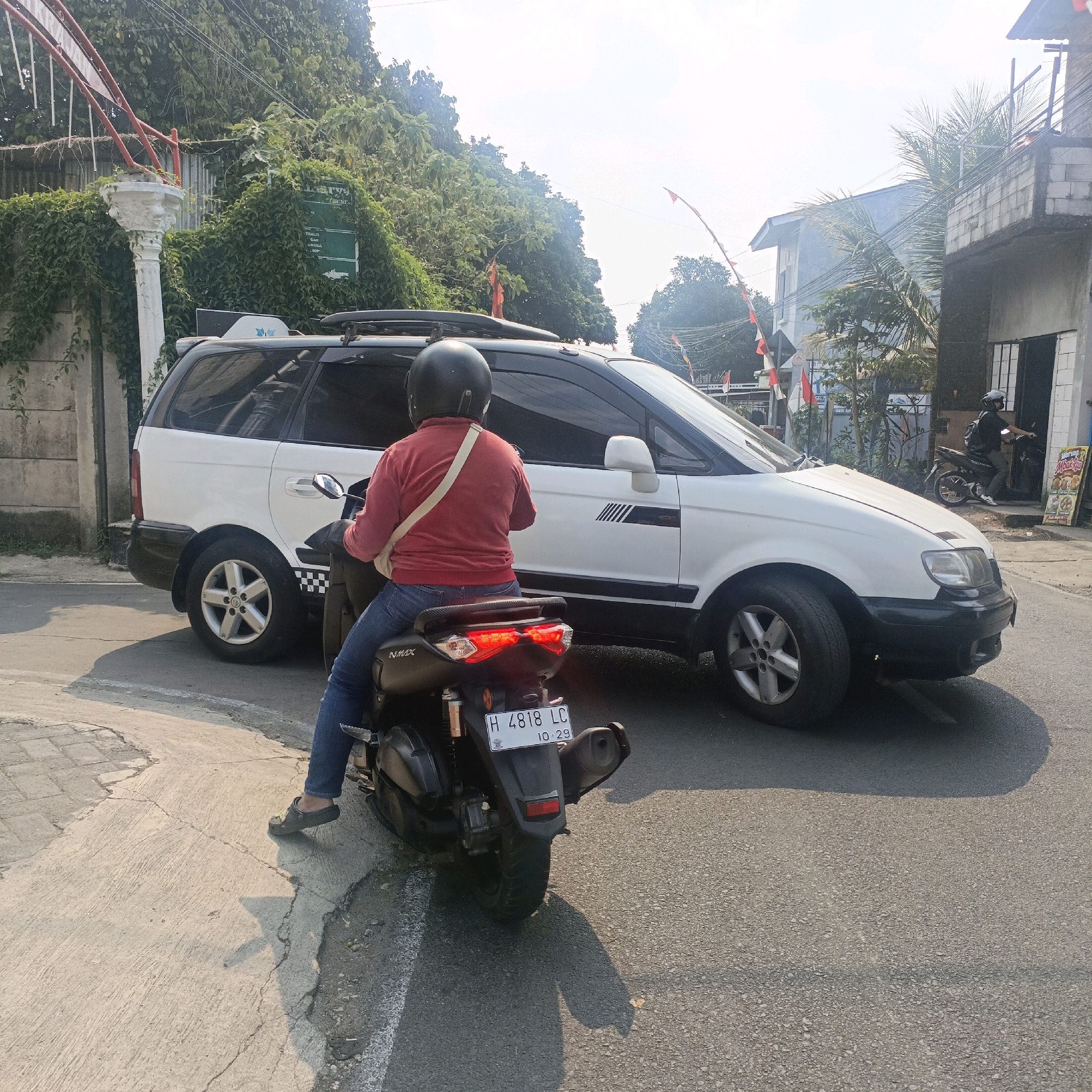 A person wearing a red long-sleeved shirt and blue jeans is sitting on a black motorcycle. The motorcycle has a license plate that reads "H 4818 LC 10.29". The person is wearing a black helmet and a light-colored bag across their chest. The motorcycle is on an asphalt road, with a cracked concrete sidewalk to its left. Behind the motorcycle, a white minivan with black trim is stopped, its rear right wheel visible. The minivan has a roof rack and appears to be a taxi or transport vehicle. In the background, lush green trees and foliage cover a hill. Several buildings, including a two-story structure with a balcony, are visible to the right, some adorned with small red and white flags, suggesting a local celebration or national holiday in Salatiga, Indonesia. The scene is illuminated by the sun, casting shadows on the road.