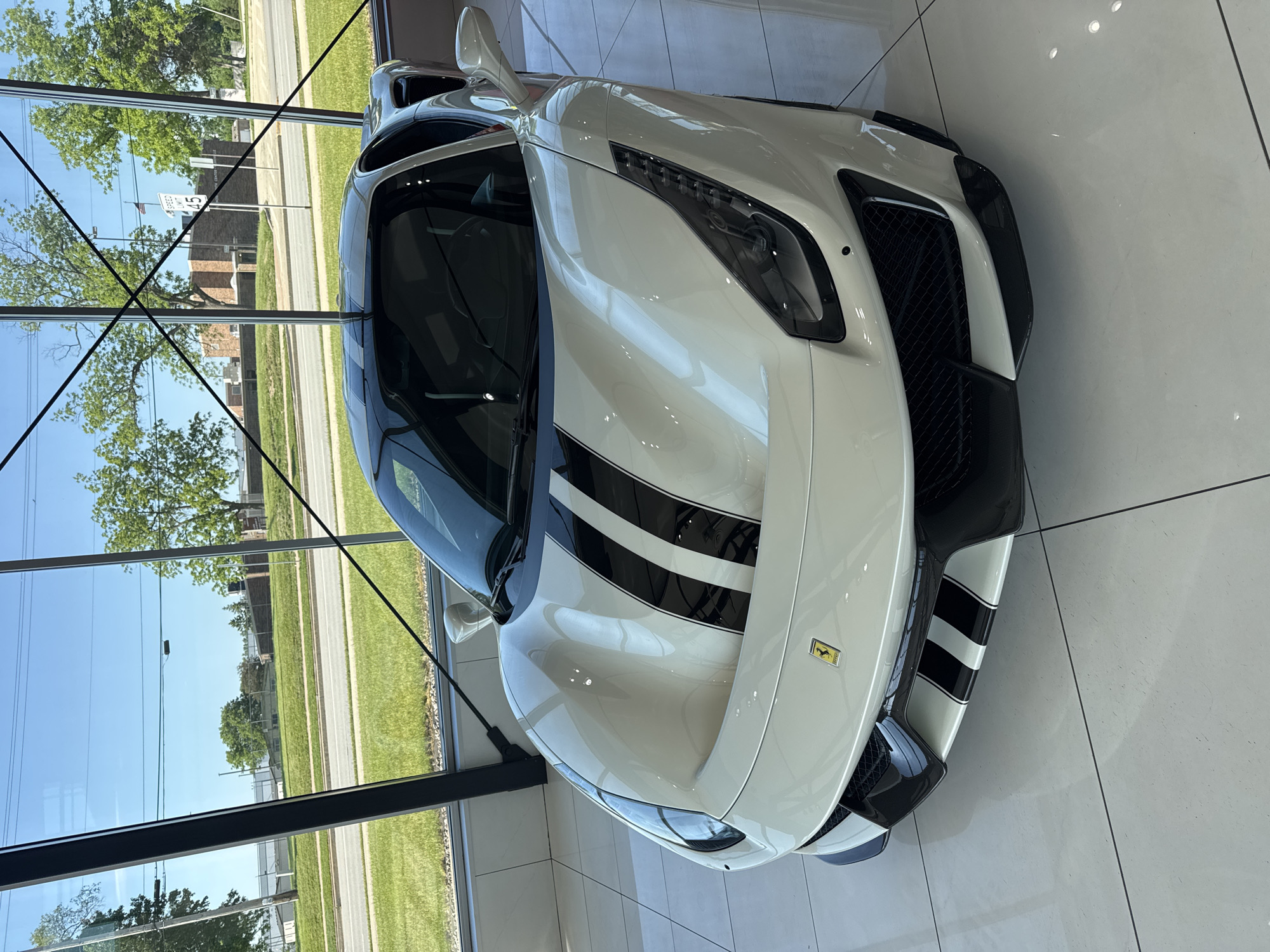 The image shows a white Ferrari sports car with black racing stripes parked inside a building with large windows. Outside the windows, a streetscape is visible. A speed limit sign indicating "45" is present. Based on the speed limit sign being visible on Ogden Avenue, the location is likely a car dealership in Naperville, Illinois, United States of America. The floor inside the building is light colored and tiled. The car is positioned at an angle, with its front facing the viewer. No people are visible in the image.