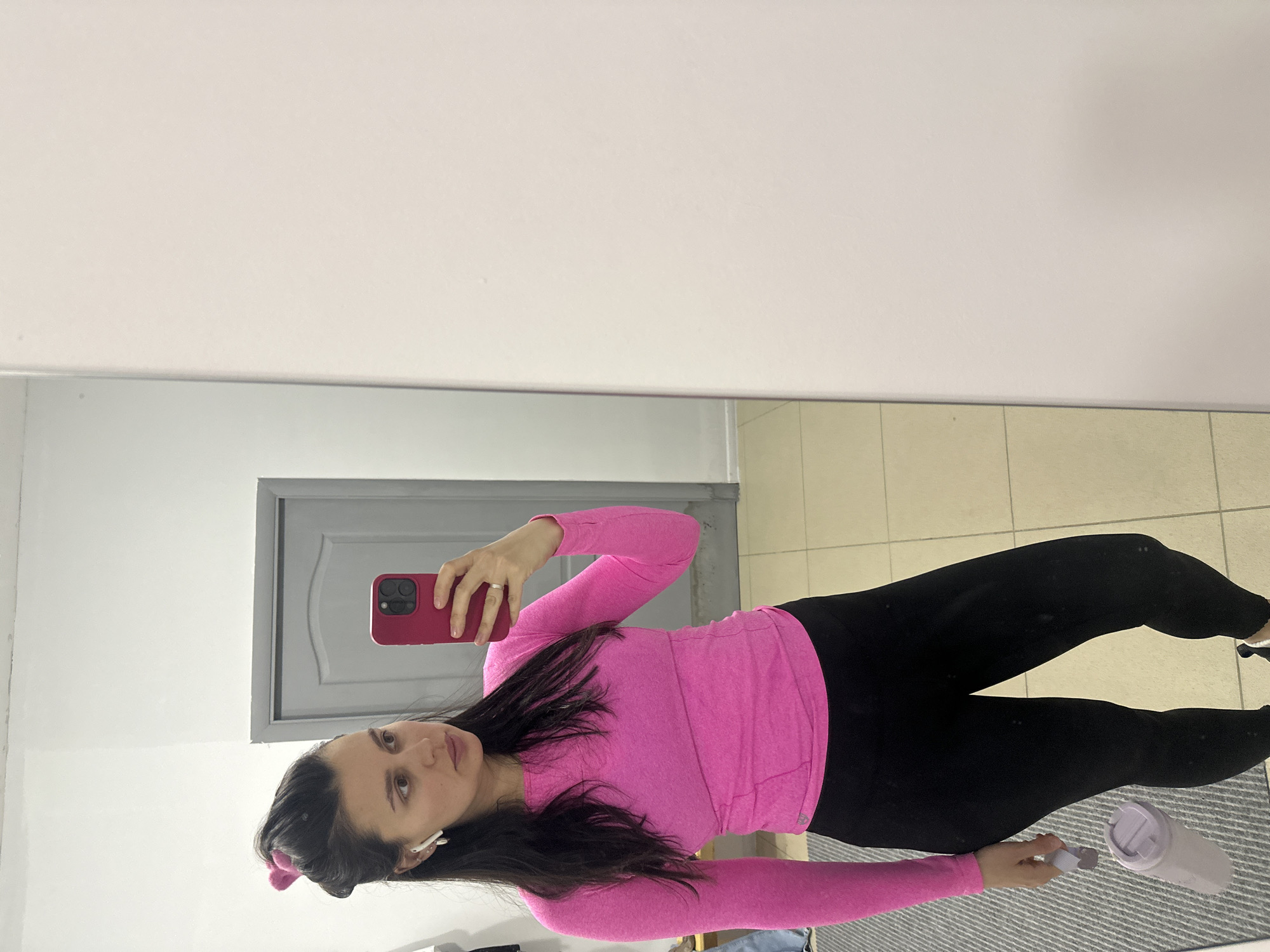 A woman is captured taking a mirror selfie in an indoor setting. She stands in front of a large mirror, holding a red-cased smartphone in her right hand, angled to photograph her reflection. Her left hand holds a light purple water bottle or shaker. She has long dark hair, parted on the side, with a pink scrunchie or hair accessory visible near her left ear. She is wearing white earbuds and has a neutral expression, looking directly at the camera.
She is dressed in a vibrant pink long-sleeved athletic top and black leggings. The immediate background visible in the mirror behind her is a plain white wall. Reflected further back in the mirror is a light grey door with a raised panel design, suggesting an adjacent room or hallway. The floor beneath her reflection is a light-colored tiled surface, partially covered by a striped grey and white rug or mat. The lighting is even, indicating an indoor environment, though the specific time of day is not discernible. The provided context indicates the location is Chaikovsky, Russia. There is no visible text in the image.