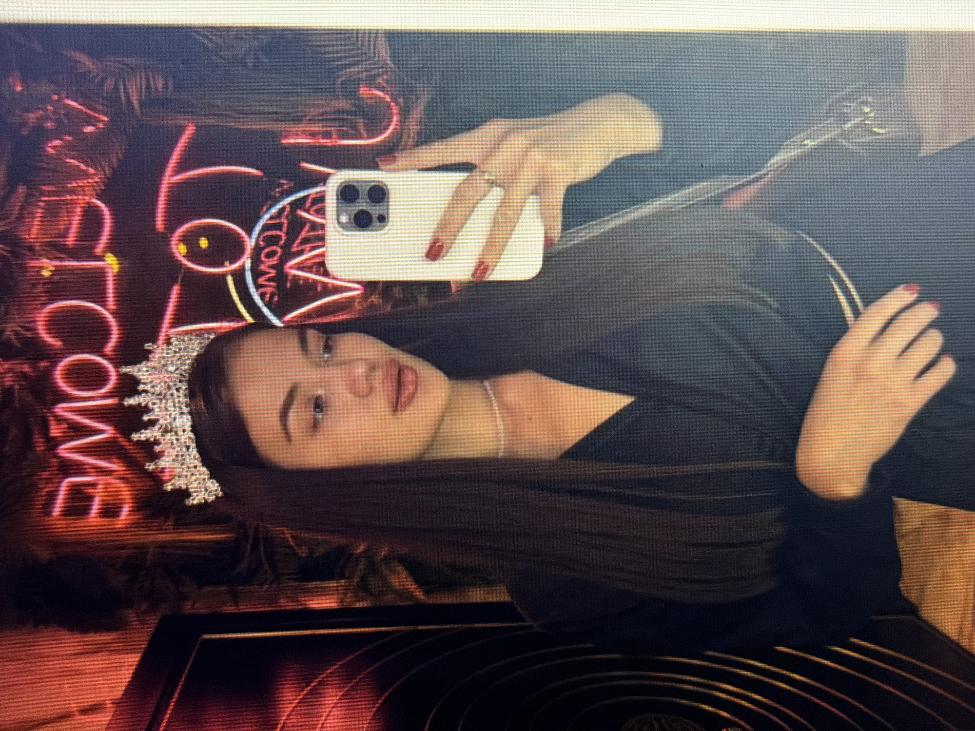 A young woman with dark hair and a tiara is taking a selfie in a dimly lit room with neon lights. She is wearing a black dress and a pearl necklace. The neon lights appear to spell out words in a foreign language, possibly Russian, and are reflected in a mirror behind her. The woman is holding a smartphone in her right hand, and her left hand is resting on her chest. The environment appears to be a nightclub or bar. The image is taken from a slightly high angle, focusing on the woman's face and upper body. The background is blurred due to the lighting and focus.