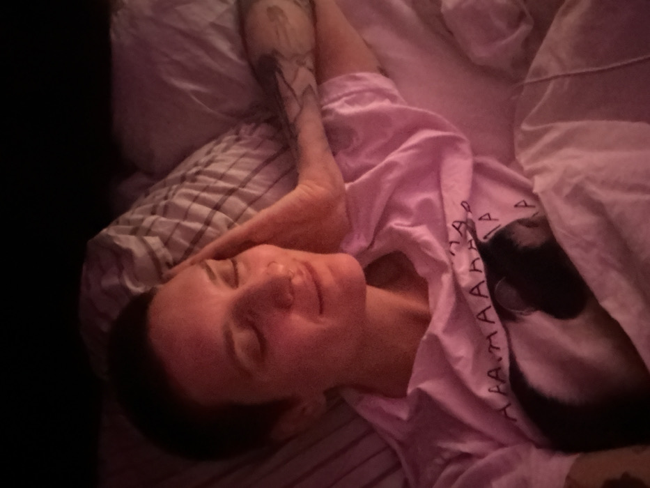 A woman with closed eyes lies in bed. She is wearing a light pink t-shirt with white vertical text. Her head rests on a striped pillow. A tattoo is visible on her raised arm. A white blanket with pink accents is draped over her. A black-and-white animal, possibly a cat, is nestled near her side, partially concealed under the blanket. The room is dimly lit.