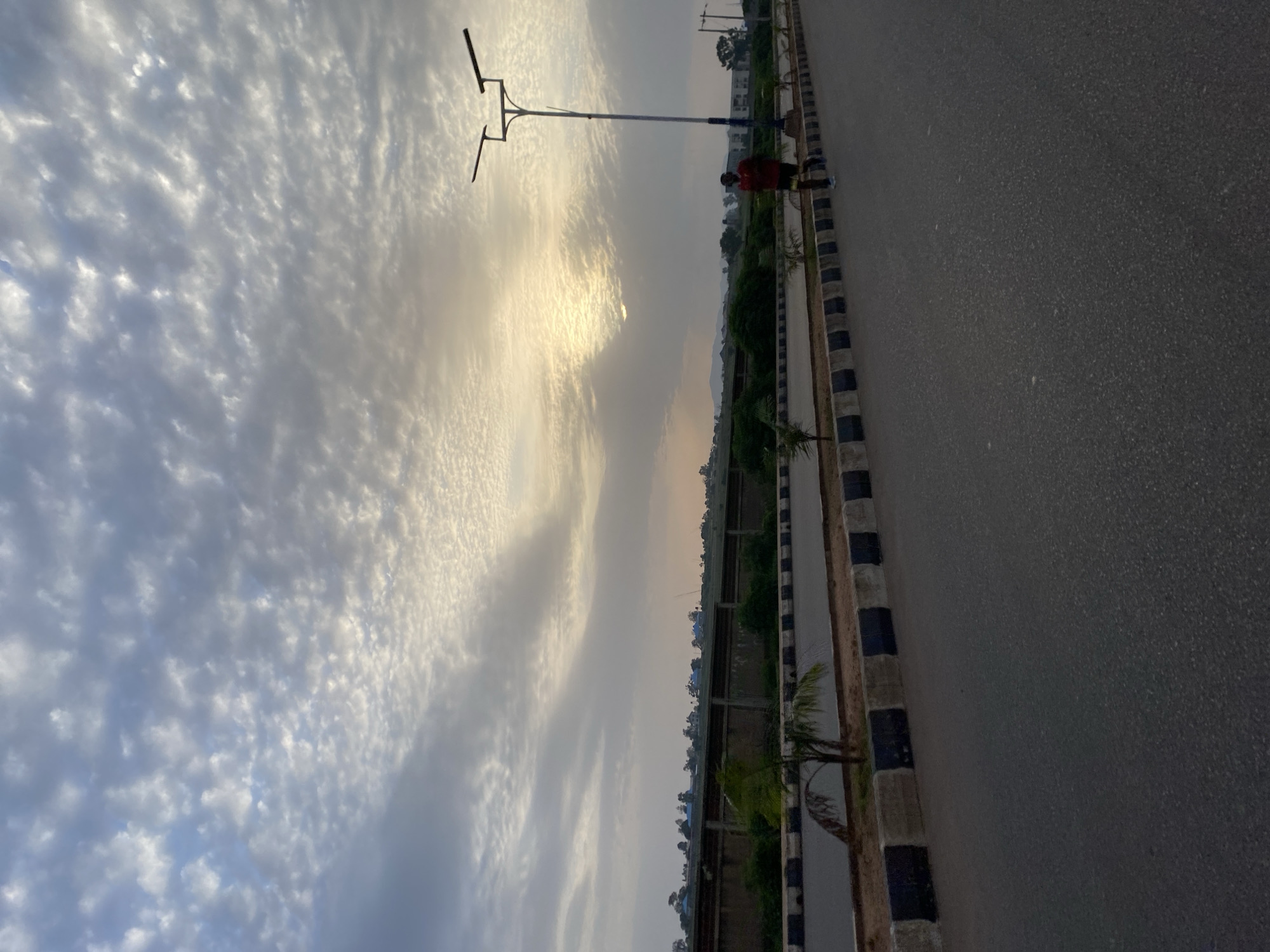 A single person is jogging on a paved road in Jos, Nigeria, under a cloudy sky with the sun peeking through. The road has a black and white striped curb. There are streetlights and small palm trees planted along the side of the road. In the background, there is a long concrete wall, vegetation, and some buildings. The sky is filled with scattered, light-colored clouds.
