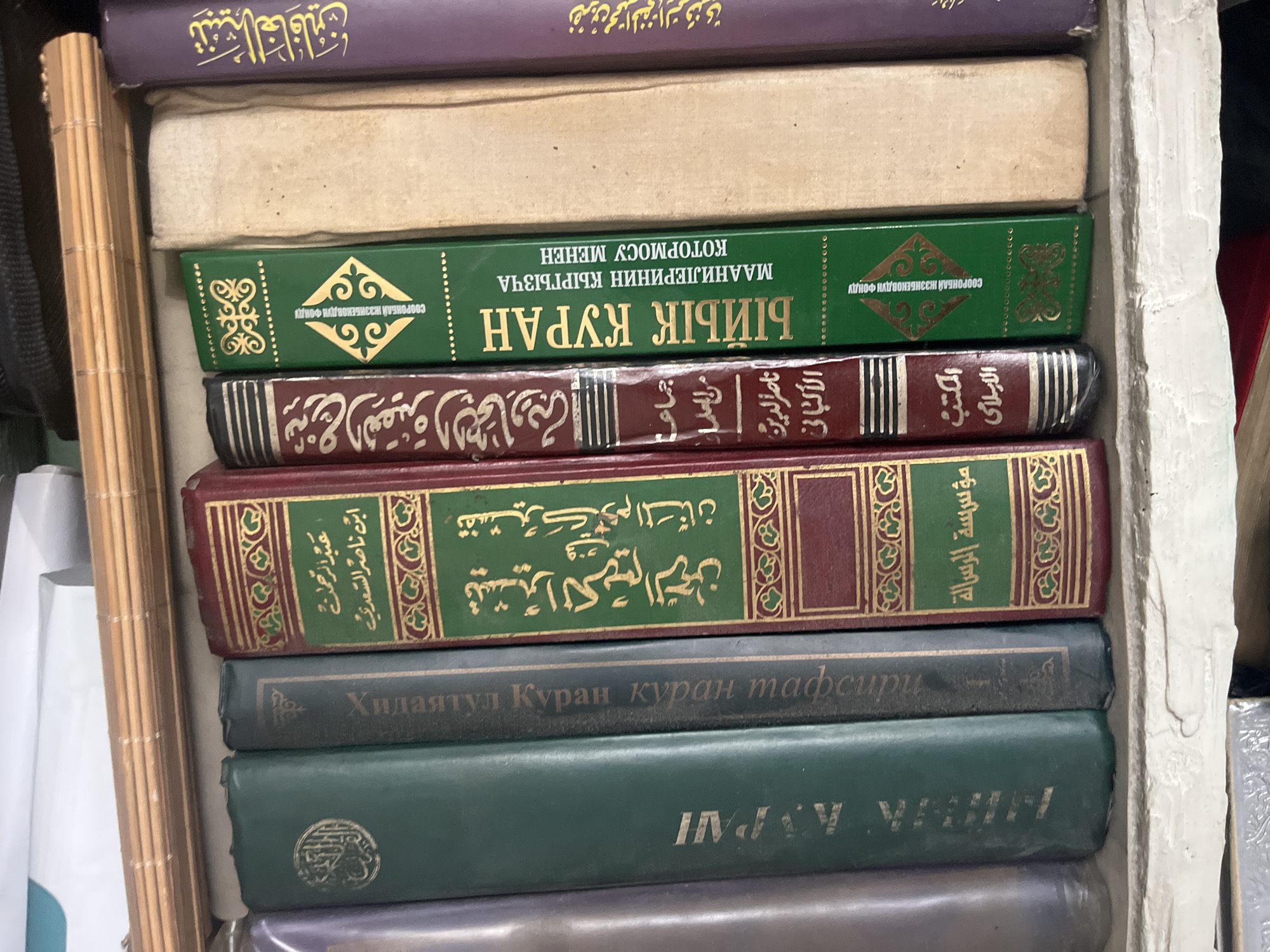 The image displays a close-up view of a shelf filled with books. The books are standing upright and appear to be of various sizes and bindings. Some of the book spines have visible text, written in what appears to be Arabic or a similar script, as well as Cyrillic script. The shelf itself is made of a light-colored, possibly concrete or plaster material, and is slightly worn. Above the books, a slatted wooden object is visible, suggesting it might be a part of a blind or a decorative element. The background is out of focus but suggests an indoor environment. No people are present in the image, and no specific location within the city of Ташлак, Kyrgyzstan, can be definitively identified from the visual information alone.
