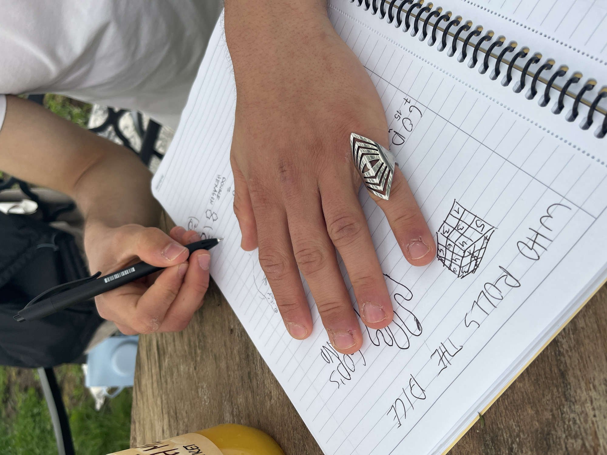 A person sits outdoors at a wooden table, writing in a spiral-bound notebook. The notebook is open to a page with the heading "WHO ROLLS THE DICE" and a sketch of a cube with numbers 1-6 written on the visible faces. The person is writing with a black pen. The person is wearing a white shirt and a distinctive silver ring on their ring finger. Part of a black backpack and a bottle of what appears to be juice are visible on the table. The location in the city of Varna, Bulgaria, is not determinable from the information given in the image.
