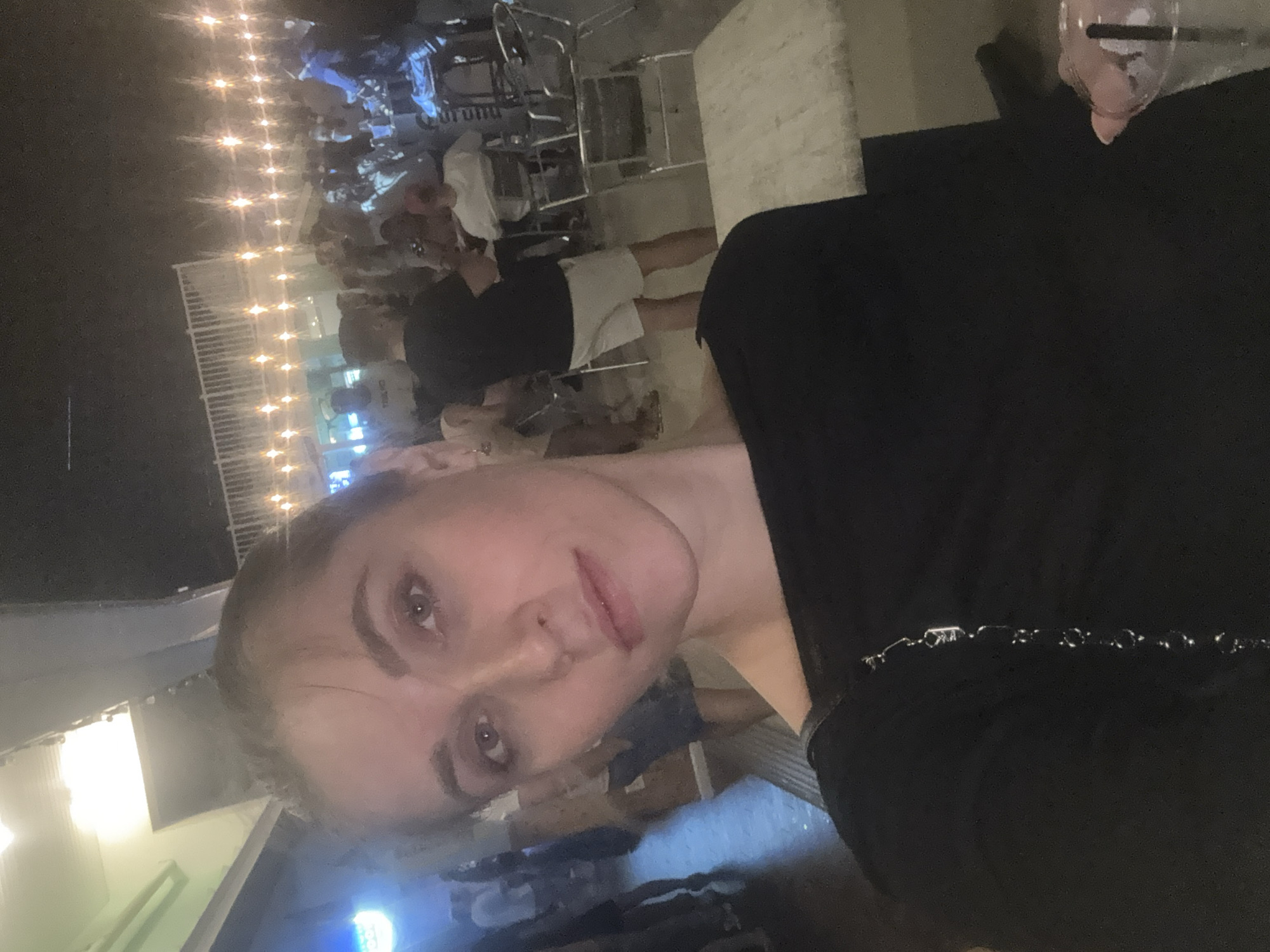 A woman is taking a selfie outdoors at night, with a crowd of people in the background. The scene appears to be a busy outdoor gathering or bar, possibly at the Jersey Shore, given the lights and the general atmosphere. The woman is wearing a black top and has her hair pulled back. The background is filled with people mingling and sitting at tables, illuminated by string lights. The location is identified as Seaside Heights, United States of America.