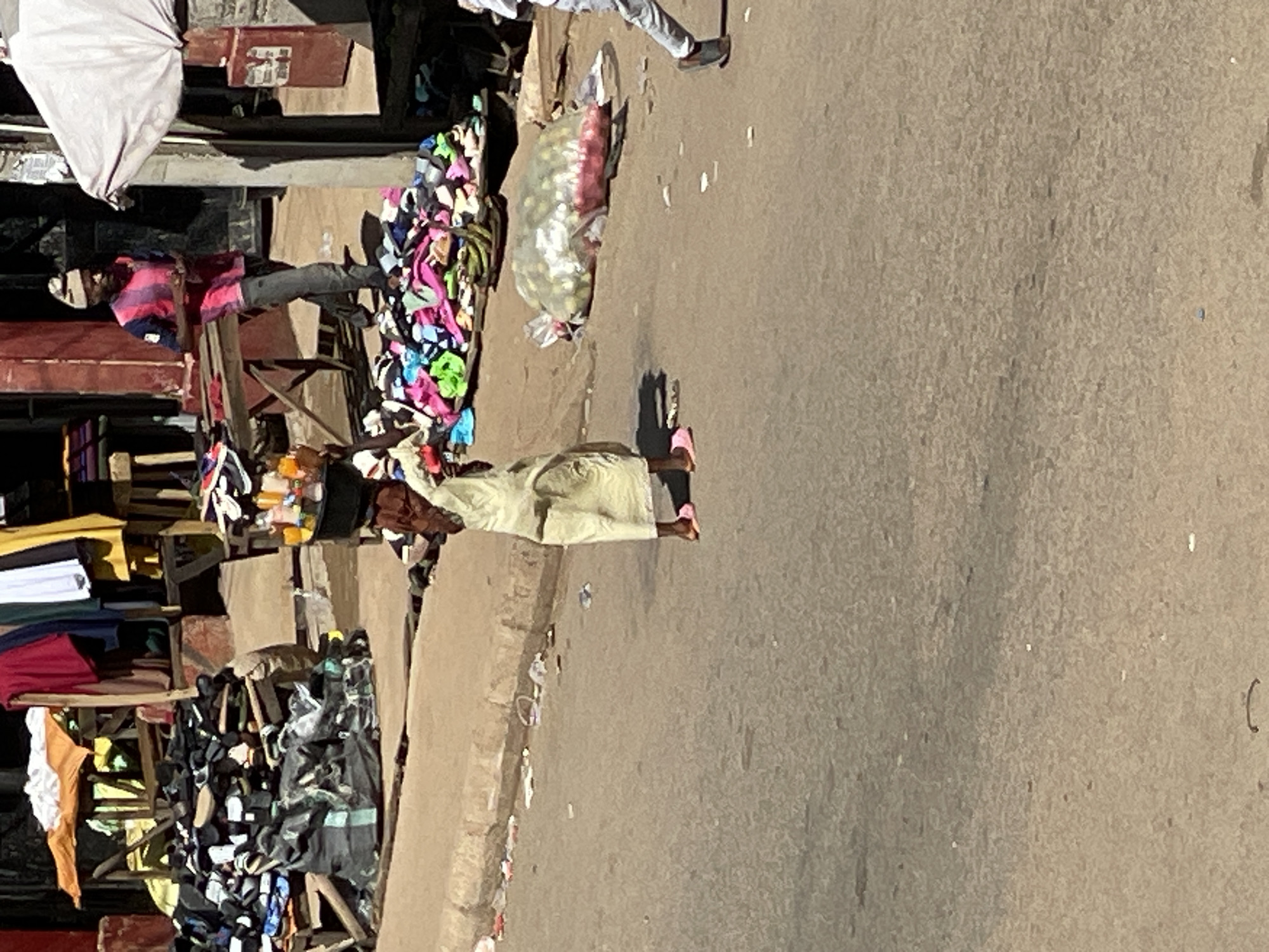 The image depicts a street scene in Bukuru, Nigeria. A woman in a light-colored dress walks along the road, away from the camera's viewpoint. Alongside the road, street vendors have set up stalls displaying various items including clothing, shoes, and other goods. A white parasol provides shade for one of the stalls. A bag filled with lemons is on the ground. Other people can be seen around the vendors' stalls.