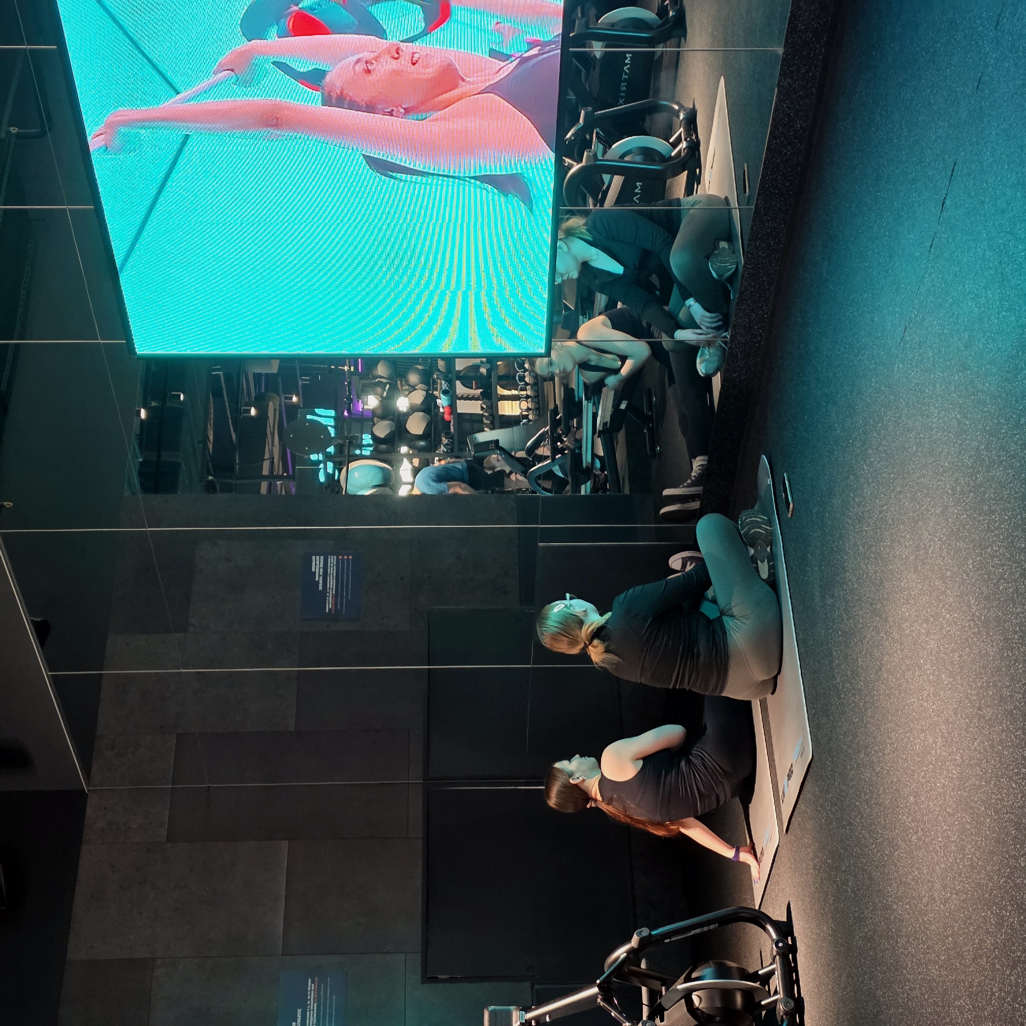 The image depicts a modern fitness studio or gym in Saint Petersburg, Russia. The setting is characterized by a dark, minimalist aesthetic with a large, vibrant LED screen providing a focal point.
In the foreground, two women are seated on light-colored exercise mats on a dark, textured floor. The woman closer to the camera has long, dark hair and wears a dark top and leggings, with a purple wristband. The second woman, positioned behind her, has lighter hair in a ponytail and is dressed in a dark long-sleeved top and grey leggings. Both appear to be resting or observing, facing towards the left of the frame.
Dominating the upper left is a large, bright teal-colored LED screen. It displays a close-up of a fair-skinned woman with light hair, engaged in a stretch or exercise pose with her arms extended. Her focused expression suggests exertion or concentration.
The left side of the studio features a highly reflective glass wall or mirror. This surface creates intricate reflections, showing more of the large screen, as well as multiple exercise bikes. The brand name "MATRIX" is clearly visible in reflection on one of the bikes. Further reflections reveal other gym equipment and the overall layout of the modern facility. On a dark wall below the screen, a blue sign with white text is partially visible, though most of its content is unreadable; the word "ATHLETIC" might be discernible.
Artificial lighting, particularly from the bright screen, casts a cool, cyan glow across parts of the room, contrasting with the dark walls and floor. The scene suggests a guided fitness session, with participants potentially following instructions from the on-screen display.