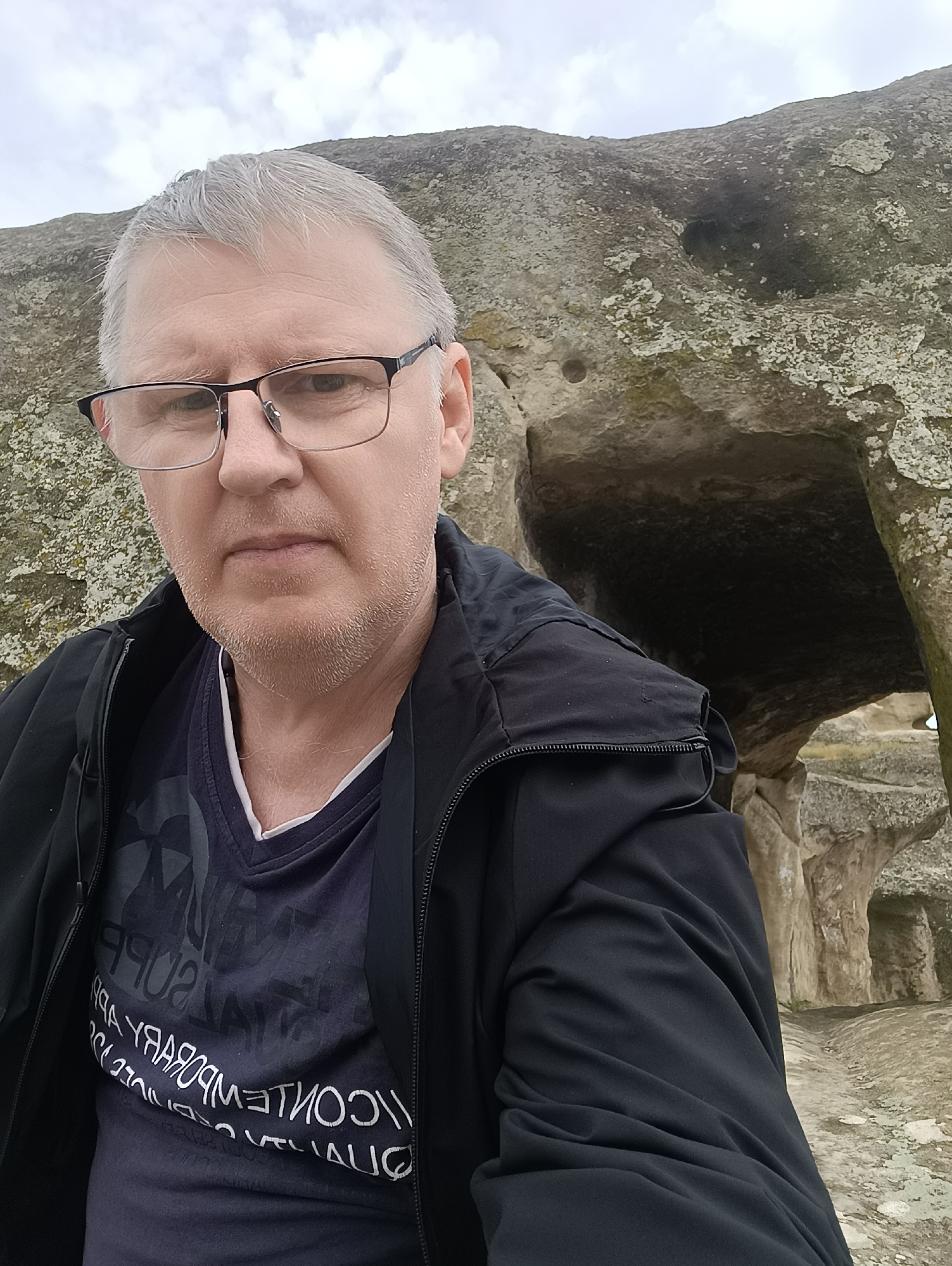 A man with graying hair and glasses is taking a selfie in front of a rock cave. He is wearing a black jacket over a dark t-shirt with white lettering. The background shows the rough, textured surface of a rock formation with an opening that appears to be a cave entrance. The sky above is overcast. The scene is likely located in Uplistsikhe, Georgia, a historic rock-hewn town.