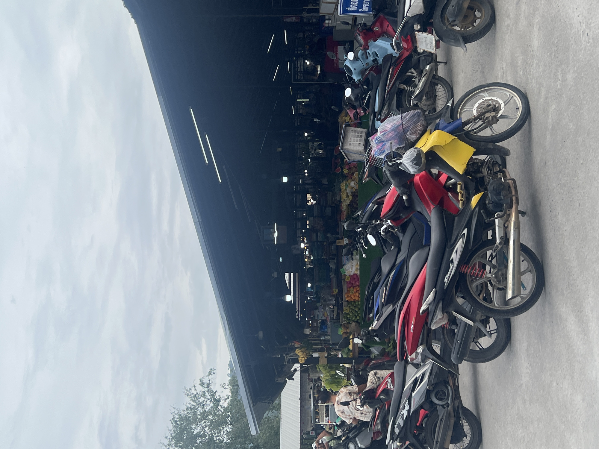 This image depicts a street market scene in the Ko Samui District of Thailand. In the foreground, several motorcycles are parked on a paved surface, with one red and yellow motorcycle prominently displayed. The motorcycles vary in color and design, suggesting a typical mode of transportation in the area. In the background, a covered market structure is visible, with stalls displaying various fruits and vegetables. A few individuals are present in the market, with one person sitting on a motorcycle and another person walking. The overall atmosphere appears to be that of a busy marketplace, with goods and people coexisting in a vibrant setting. The sky above is overcast, indicating a cloudy day.