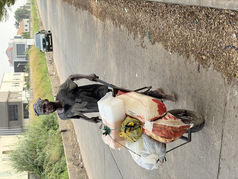 A man pulls a loaded cart on a paved road. He wears a dark shirt and head covering. The cart is filled with various bags and containers, including rice. Houses and greenery are visible on the sides of the road. A car drives in the background. Dry leaves litter the edge of the road.