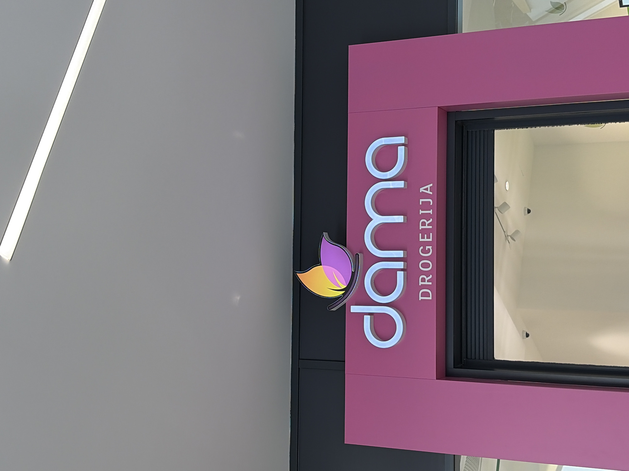 The image shows a storefront for "dama DROGERIJA," a cosmetics and drugstore. The sign is pink with white lettering and a butterfly logo that has yellow, orange, and purple hues. The store is in Budva, Montenegro. A window is visible with a black frame and displays items inside. A bright white light is present on the ceiling.