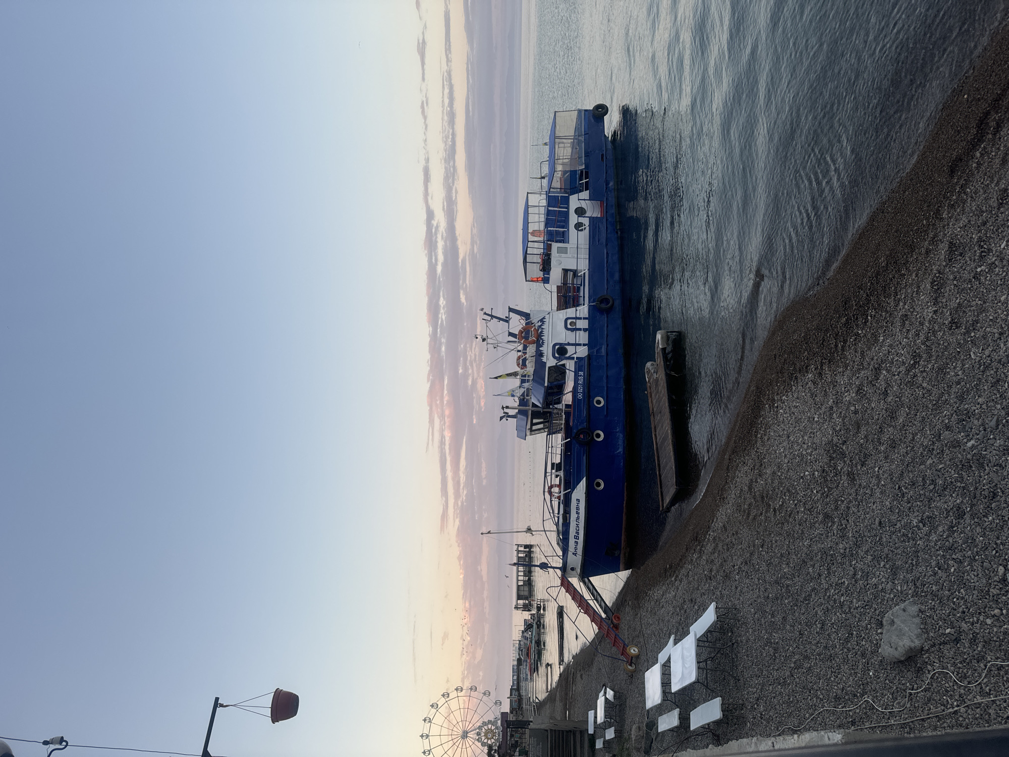 In Listvyanka, Russia, on a pebbled shoreline, a blue and white ferry boat named "Anna Vasilievna" is docked. The boat has a visible hull number "OO 0251 RUS 38." A red and orange ramp with wheels extends from the shoreline to the boat's entrance. Several white picnic tables and benches are arranged on the shore near the ramp. In the background, a Ferris wheel and other structures line the waterfront. The sky displays a gradient of soft blues and pinks, indicating either sunrise or sunset, with scattered clouds. The calm water of the lake gently laps the shore.