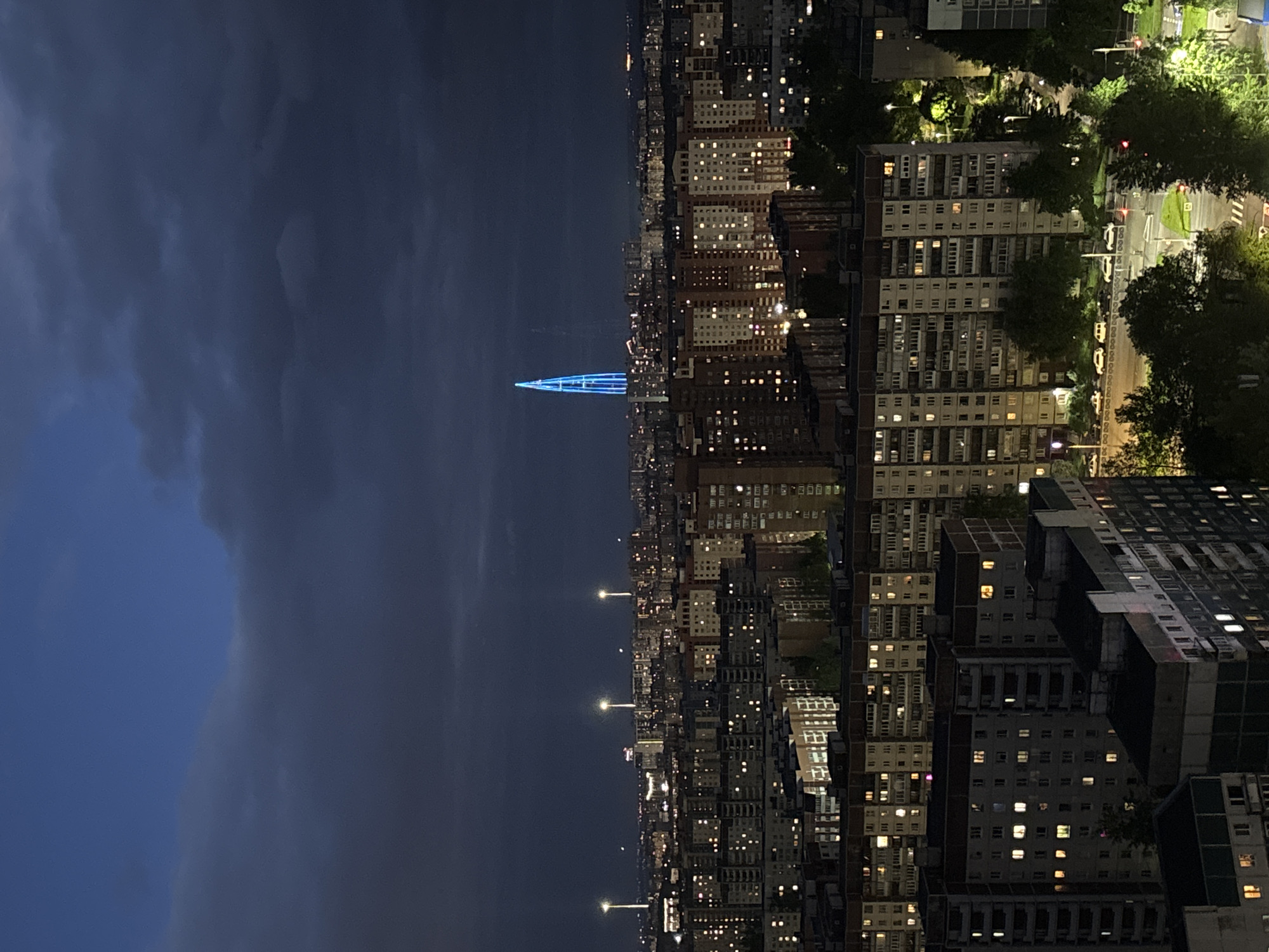 The image depicts an aerial view of Saint Petersburg, Russia, at night. The city skyline is dominated by tall apartment buildings, many with illuminated windows. Streetlights are visible along the streets. In the distance, a blue, triangular light structure pierces the dark sky, likely a landmark or architectural feature of the city. The sky is overcast with dark clouds.