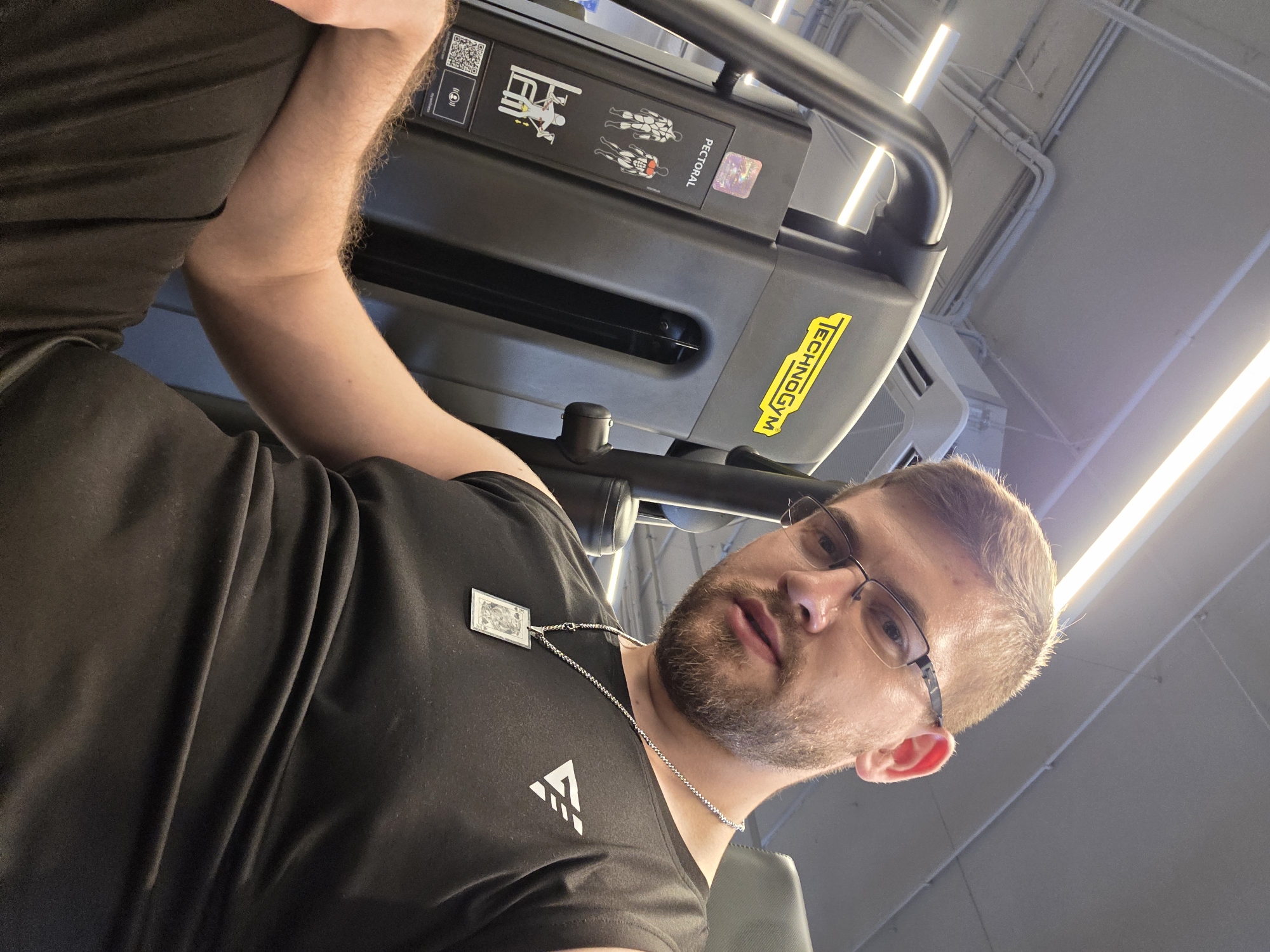 A man wearing glasses, a black shirt with a white logo, and a silver chain with a pendant is sitting on a Technogym pectoral machine. He has short brown hair and a beard. The background is a gym setting with bright overhead lighting. The location appears to be a modern fitness center, possibly in Plovdiv, Bulgaria.
