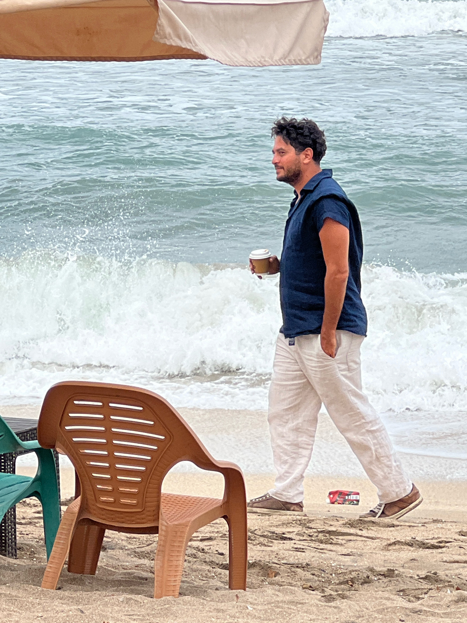 An adult male with dark, curly hair and a beard walks along a sandy beach, holding a disposable coffee cup in his right hand. He is wearing a sleeveless dark blue shirt and loose-fitting light-colored linen pants. The man is walking towards the right side of the frame, looking forward. Behind him, the ocean waves crash onto the shore, creating white spray. In the foreground, a brown plastic chair and a portion of a green chair are visible, placed on the sand. A small red and white package lies on the sand near the man's feet. An umbrella provides shade in the upper left portion of the image. The scene appears to be the beach in Kuta, Indonesia.