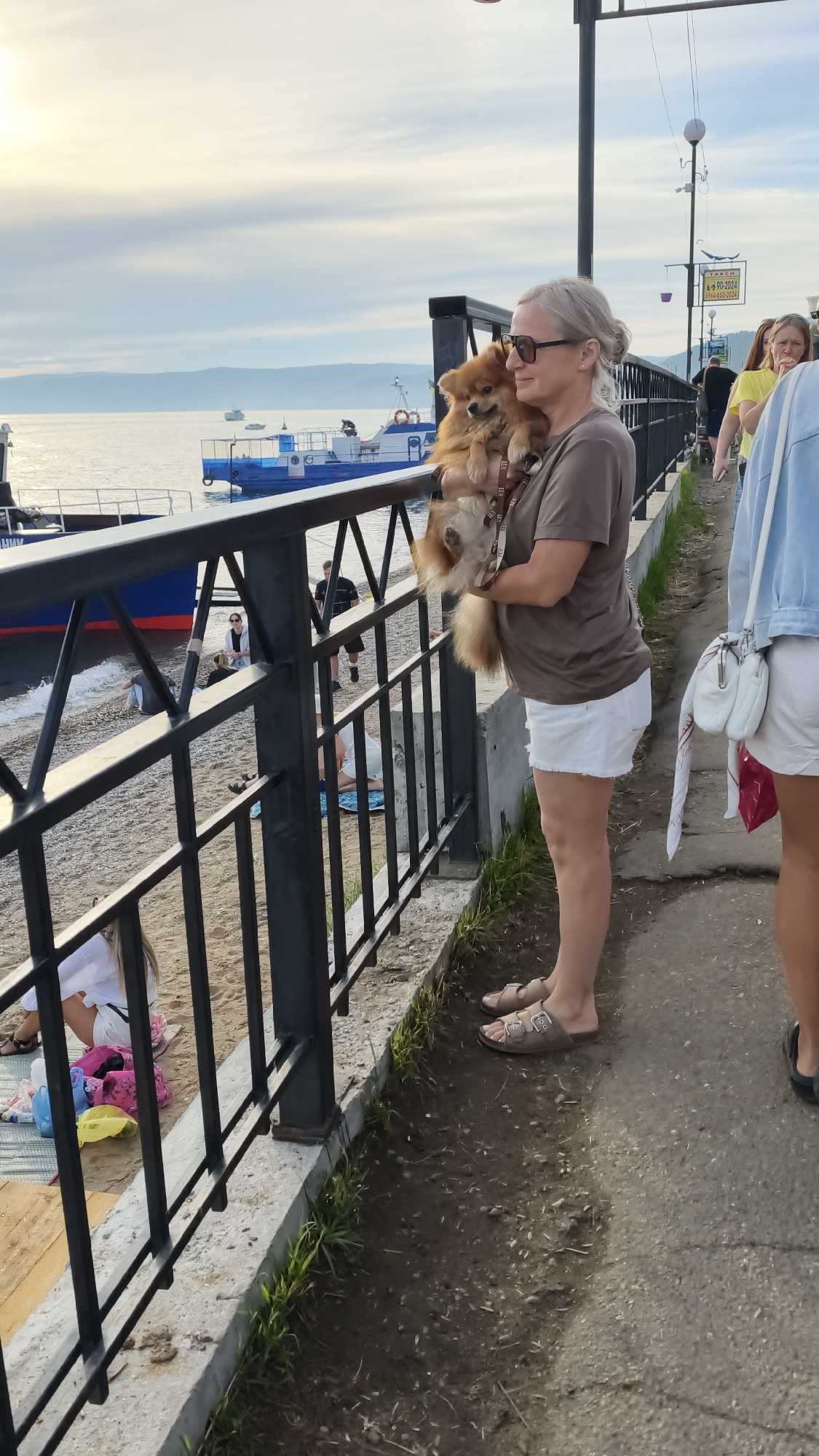 A blonde woman with sunglasses is holding a small, fluffy tan dog. She is wearing a brown t-shirt and white shorts. She is standing on a concrete walkway next to a black metal fence. Behind the fence, there is a beach area with people sitting on blankets and a body of water with boats. The location is the Listvyanka embankment in Listvyanka, Russia. The sky is overcast, and the lighting suggests it is daytime.