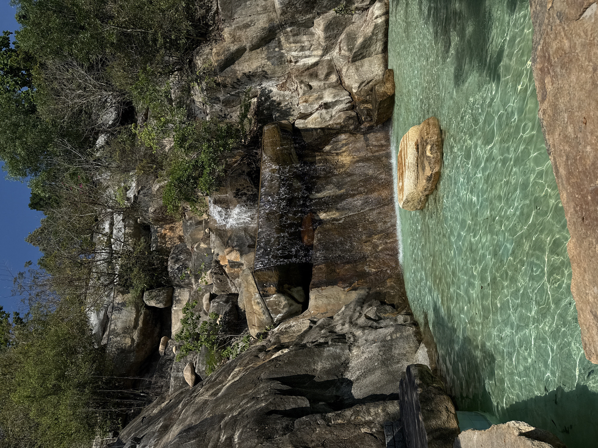 A partially obscured waterfall flows over a rocky cliff into a pool of green-tinted water, set against a backdrop of trees and a blue sky. The location is in the city of Tay Nha Trang Ward, Vietnam. Rocks and trees cover the falls and surrounding cliffs. Sunlight reflects on the water.