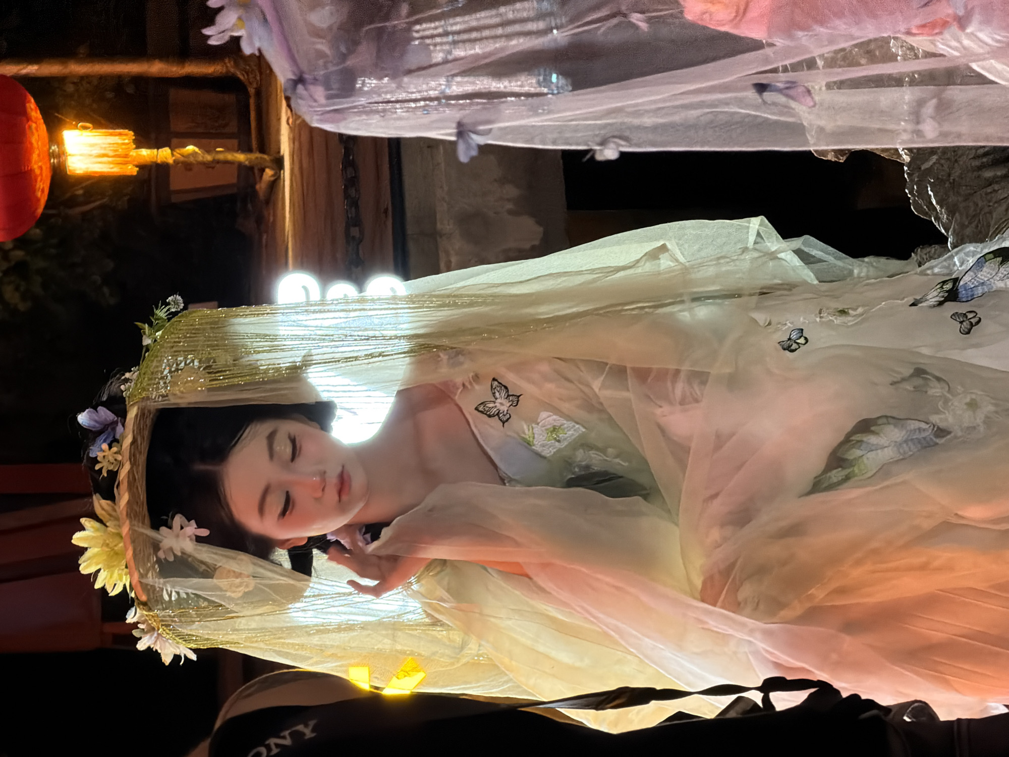 A young Asian woman is seated, wearing a pale yellow, sheer gown adorned with embroidered butterflies and floral motifs. She has a delicate headdress made of straw, decorated with yellow and purple flowers, and a gold beaded veil. Her makeup is subtle with pink blush and eyeshadow. She is touching her face with her right hand. To her left, a person in a black garment with "SONY" visible is partially visible. To her right, a sheer, pale pink veil with purple butterflies and sequins is draped. The background is dark, with a bright red lantern illuminating a wooden structure to the right. The setting appears to be outdoors at night, possibly in a park or garden area. The image is taken from a medium shot, focusing on the woman and her attire. The location is identified as Yuzhong District, China.