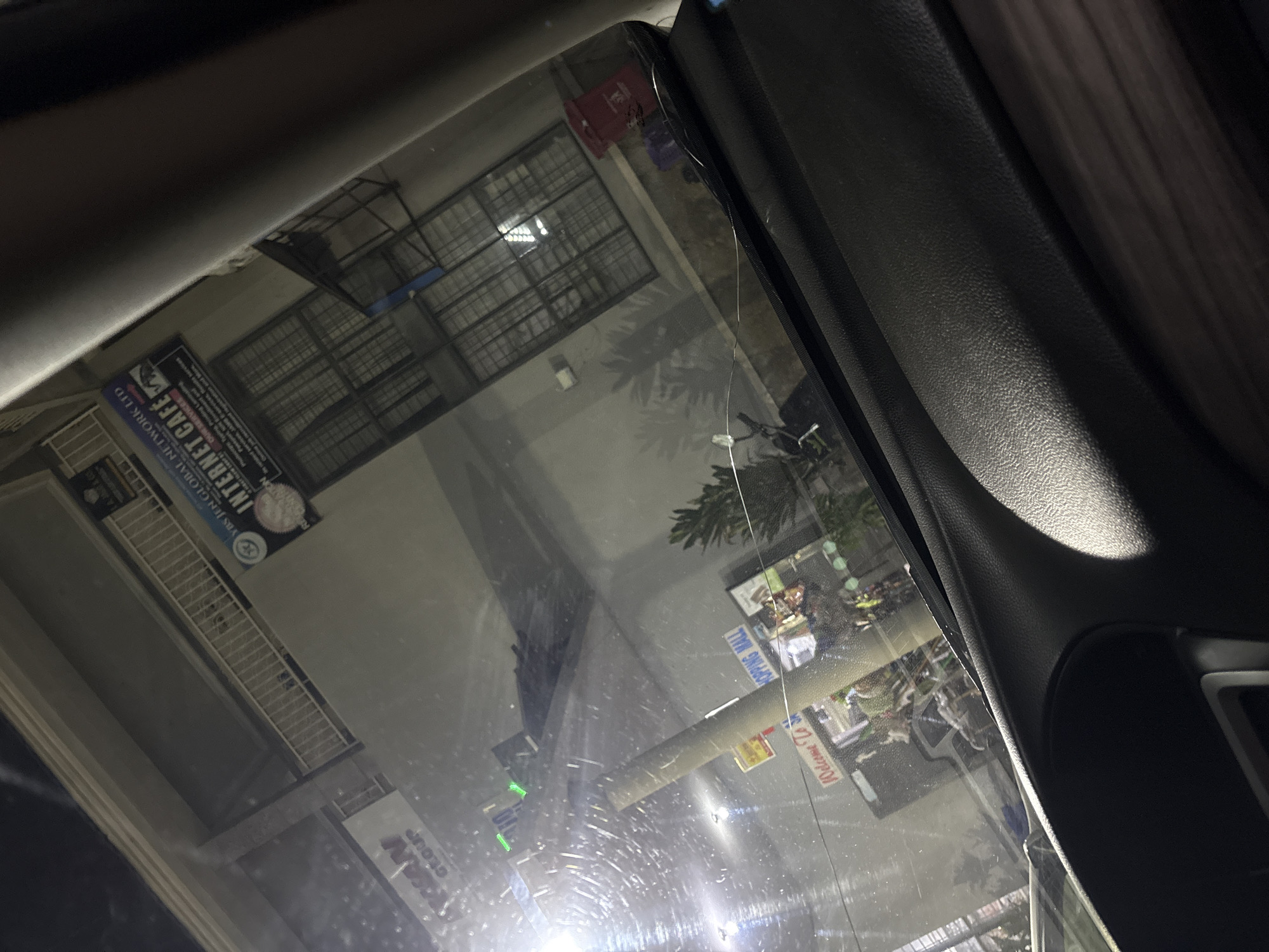 The image, taken from inside a vehicle, shows a view through the windshield at night. In the background, a building displays signage for "GLOBAL NETWORK LTD" and "INTERNET CAFE." The cafe offers services like printing, typing, browsing, and more. A "SHOPPING MALL" sign is also visible. The scene suggests an urban environment in Jalingo, Nigeria, with commercial activity illuminated at night. There are no visible people or specific actions taking place. The windshield has several streaks and smudges, and a crack is visible across it.