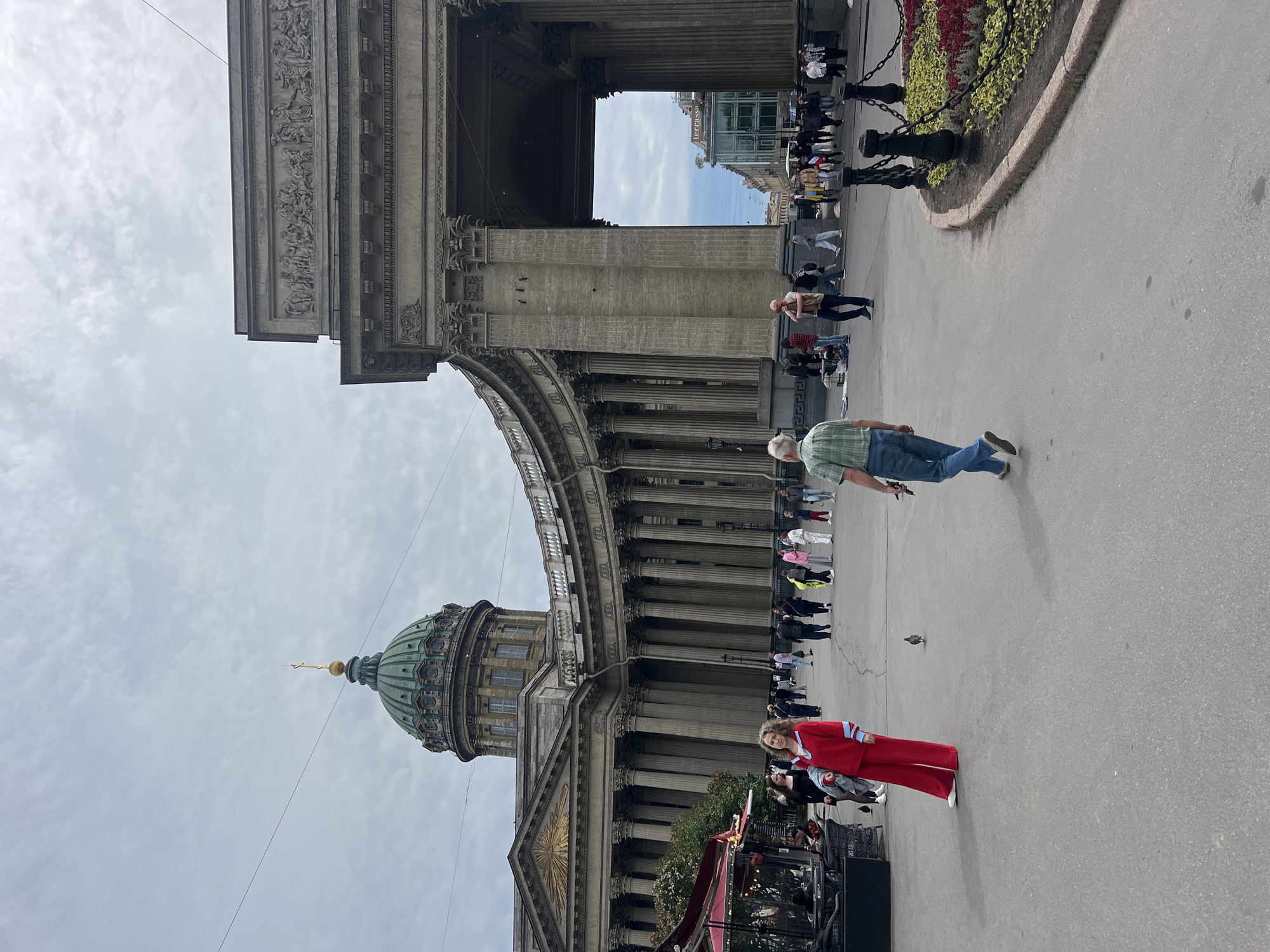 The image depicts the Kazan Cathedral in Saint Petersburg, Russia, a prominent landmark in the city. The cathedral's imposing neoclassical facade, characterized by a semicircular colonnade, is visible in the background. A grand archway, part of the cathedral's architectural ensemble, is also featured. Several people are walking in the open area in front of the cathedral, with a woman in a red suit standing prominently on the left. A man in a plaid shirt and jeans walks away from the camera in the foreground. The sky is overcast, and the scene appears to be a public gathering space or promenade.